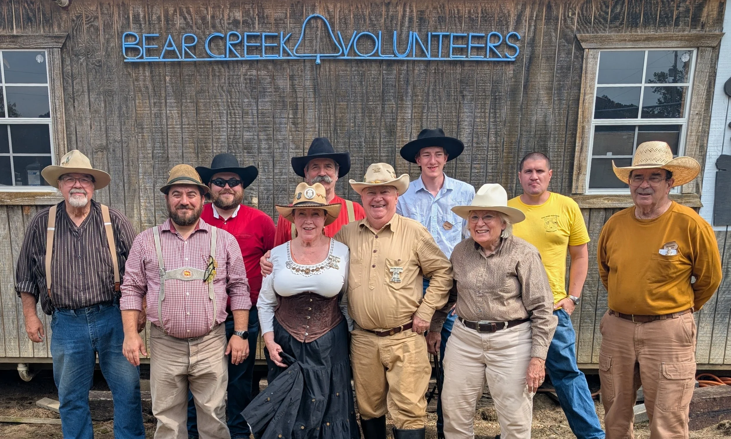 Group of people in cowboy attire standing outdoors in front of a wooden building with a sign that reads 'Bear Creek Volunteers' in blue neon letters.