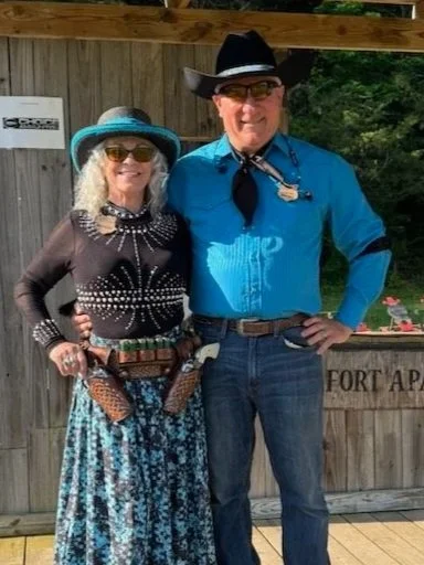A woman and a man dressed in Western-style clothing, standing outdoors in front of a wooden structure. The woman wears a wide-brimmed blue hat, sunglasses, a black top with white patterns, and a long patterned skirt, while the man wears a black cowboy hat, glasses, a blue shirt, jeans, and a bolo tie.
