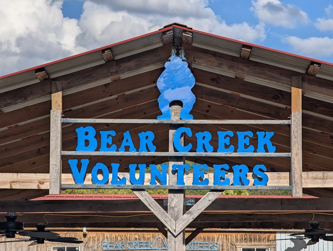 Sign for Bear Creek Volunteers with a bear figure above the text, located on a wooden structure with a metal roof, under a partly cloudy sky.