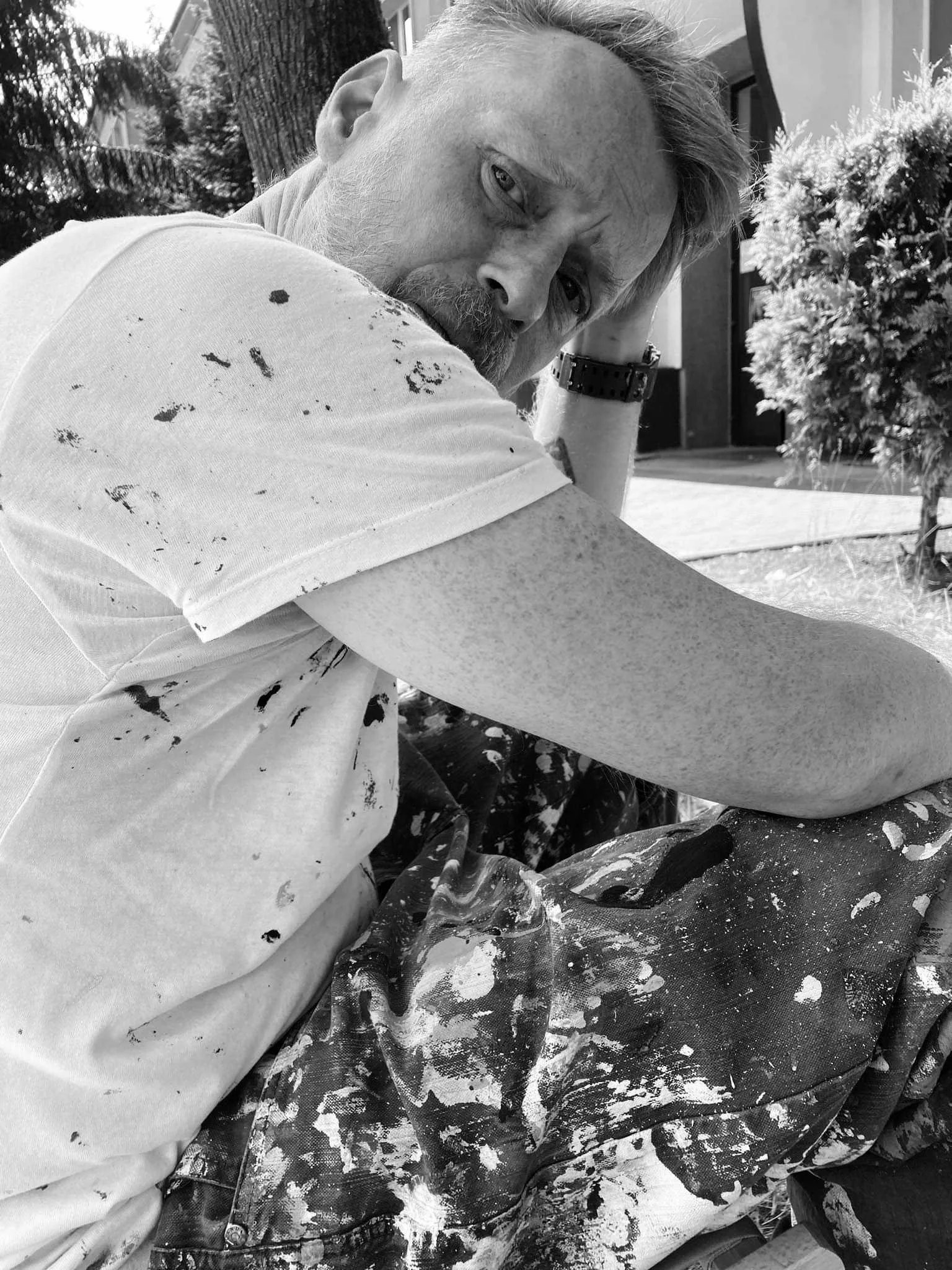 A man with a beard and blond hair, wearing a white t-shirt with paint splatters, sitting outdoors on a sunny day, resting his head on his hand and looking at the camera.