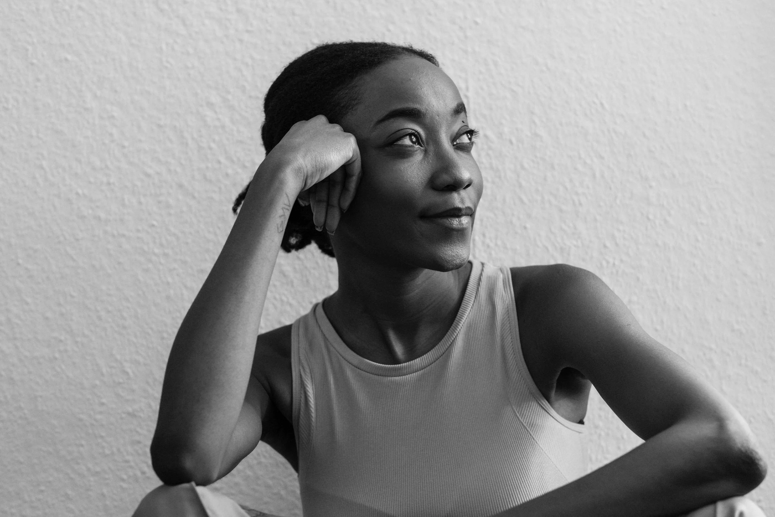 Black and white photo of a woman with braided hair, sitting with her arm resting on her knee, looking thoughtfully to the side, wearing a sleeveless top.
