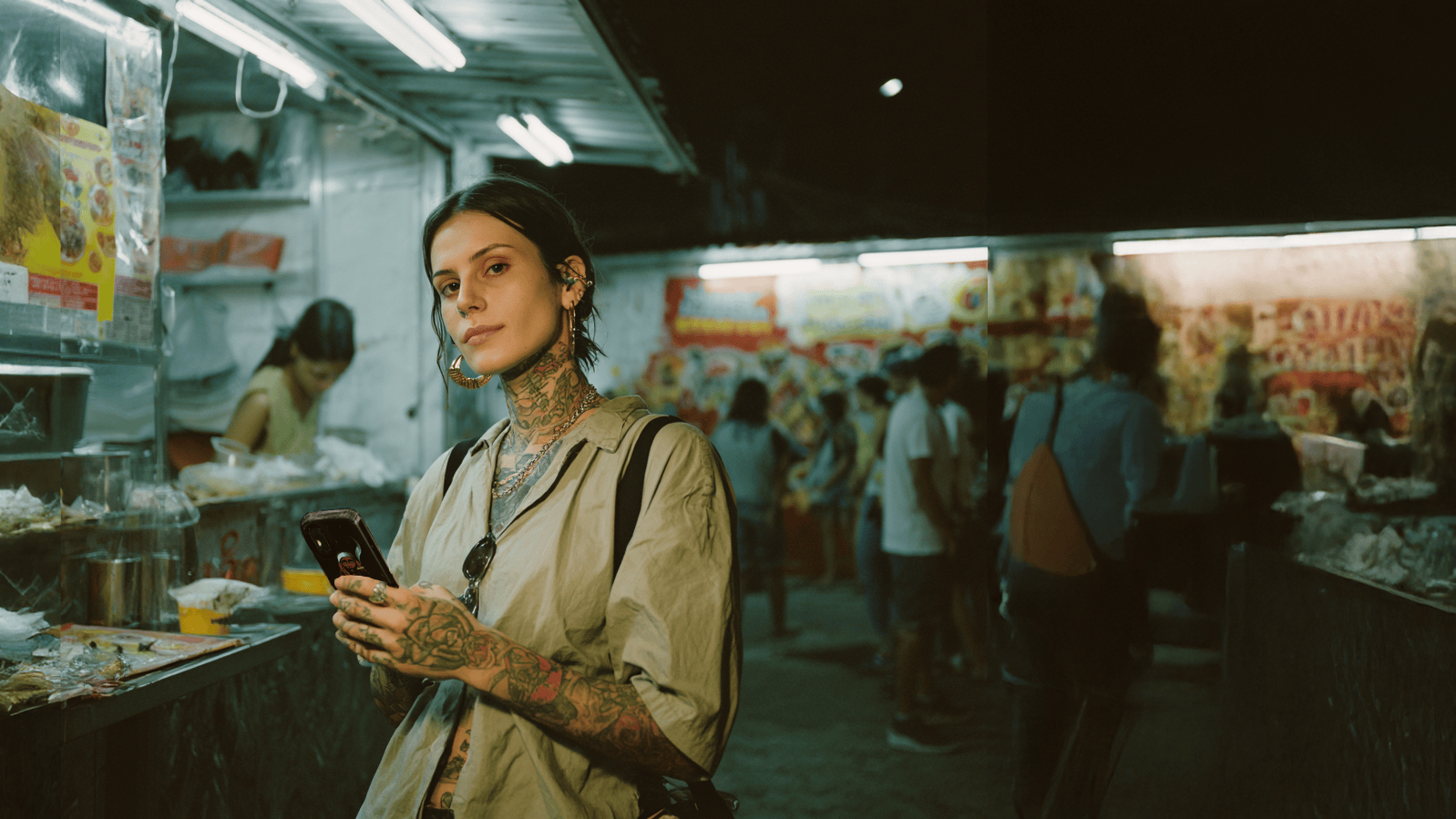 Young woman with tattoos on her neck and arms holding a smartphone in a nighttime outdoor market
