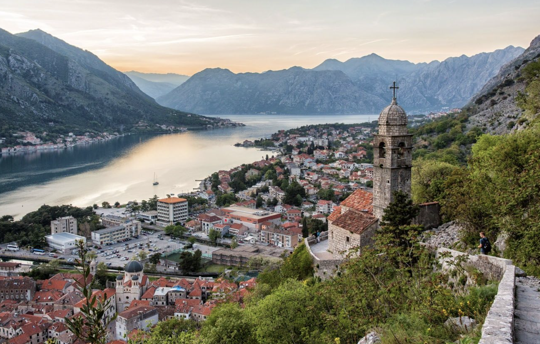 A scenic view of a coastal town with a river or bay, surrounded by mountains, during sunset. The town has historic buildings and a church with a bell tower. There are trees and a hillside walkway in the foreground, and a person is seen walking along the path.