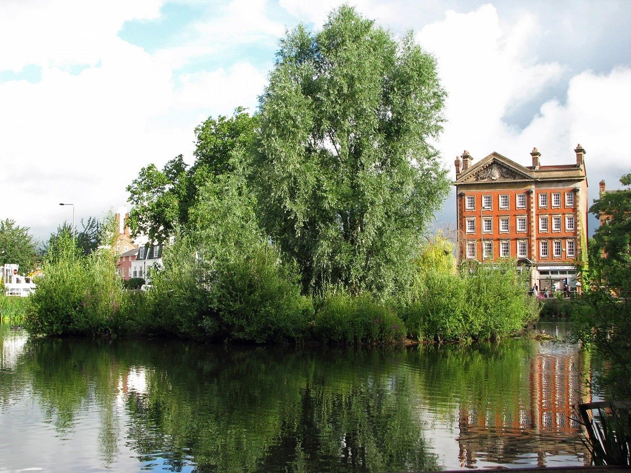 A serene riverside scene with trees and shrubs reflected in calm water, and a red brick building with multiple windows in the background.