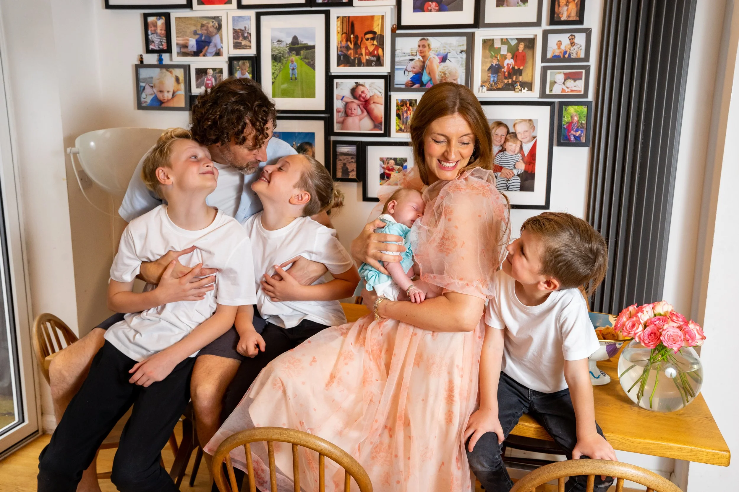 A family gathering in a room decorated with numerous photos on the wall. A woman holding a baby is smiling and surrounded by five children who are interacting affectionately with each other and the woman.