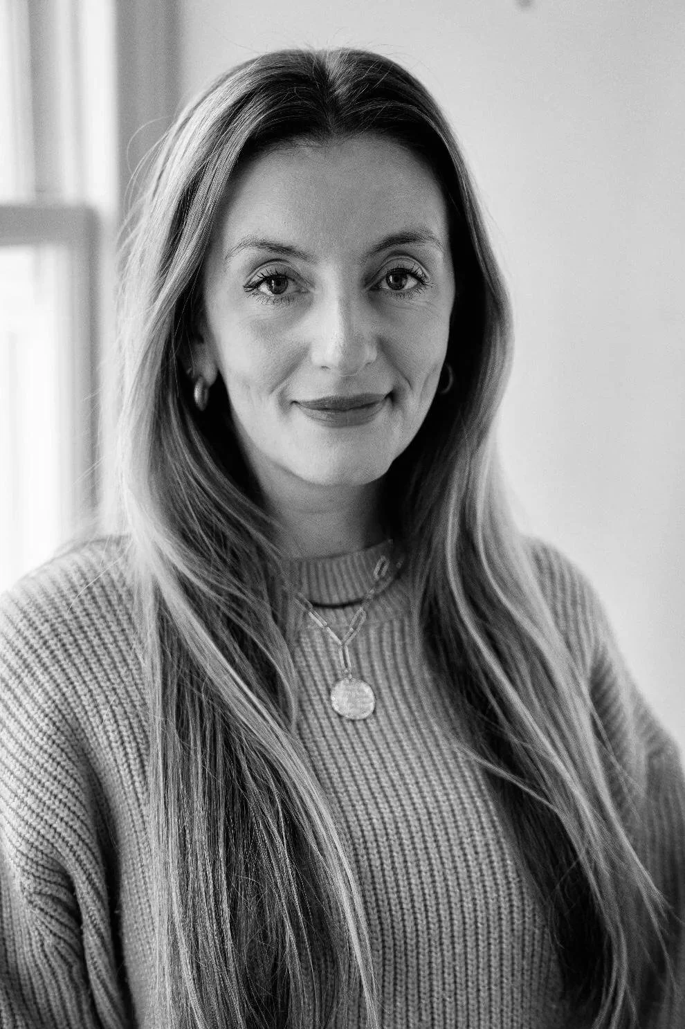 A woman with long hair and earrings, wearing a striped sweater, standing near a window in a room.