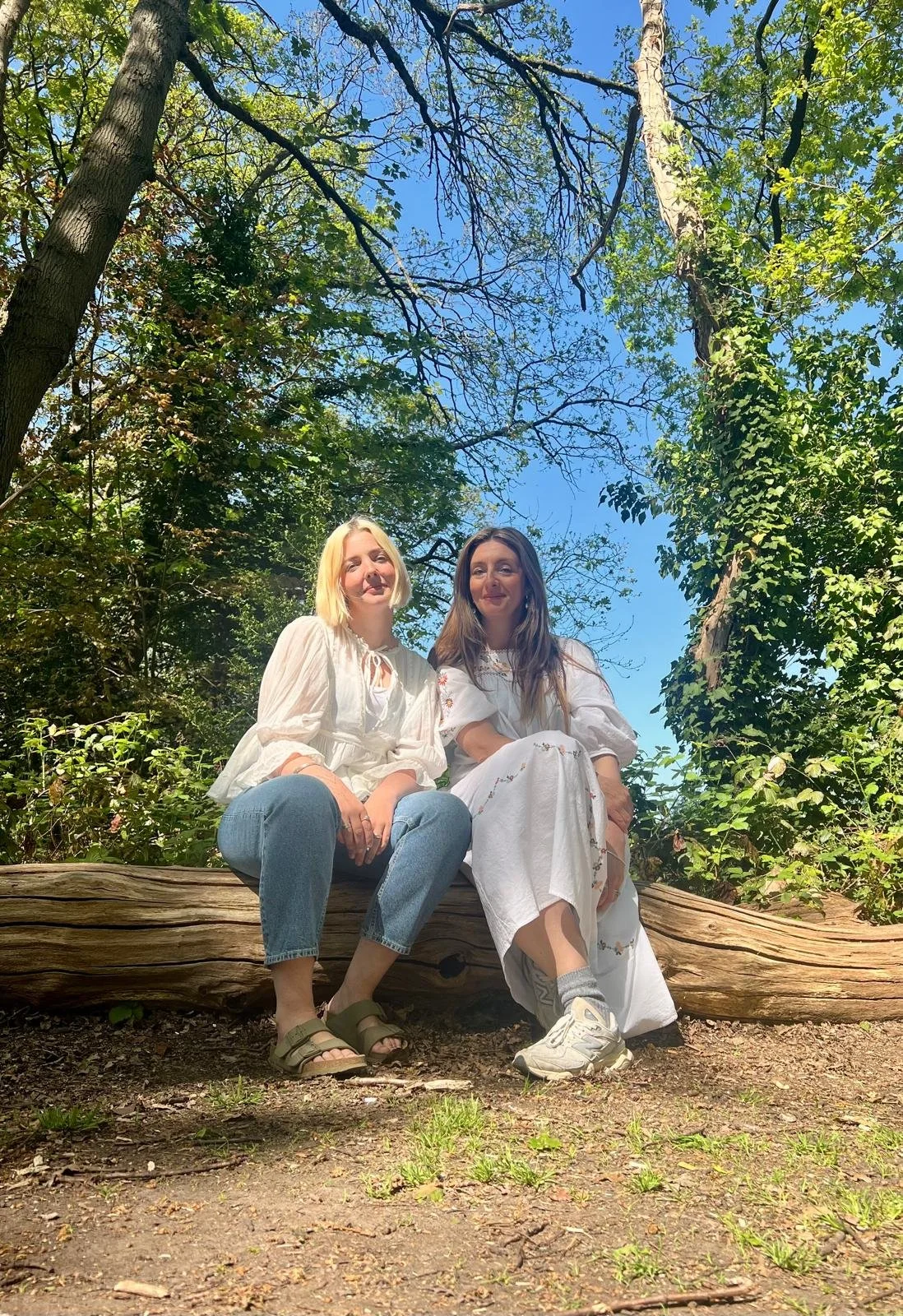 Two women sitting on a large fallen tree trunk in a forest with tall trees and green foliage, under a clear blue sky.