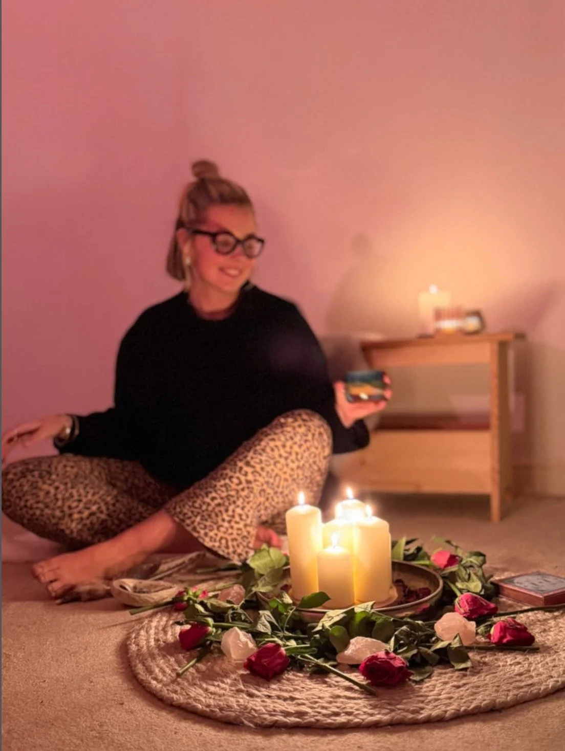 Woman sitting cross-legged on carpet, surrounded by candles and roses, holding a phone, with a desk and lit candle in the background.