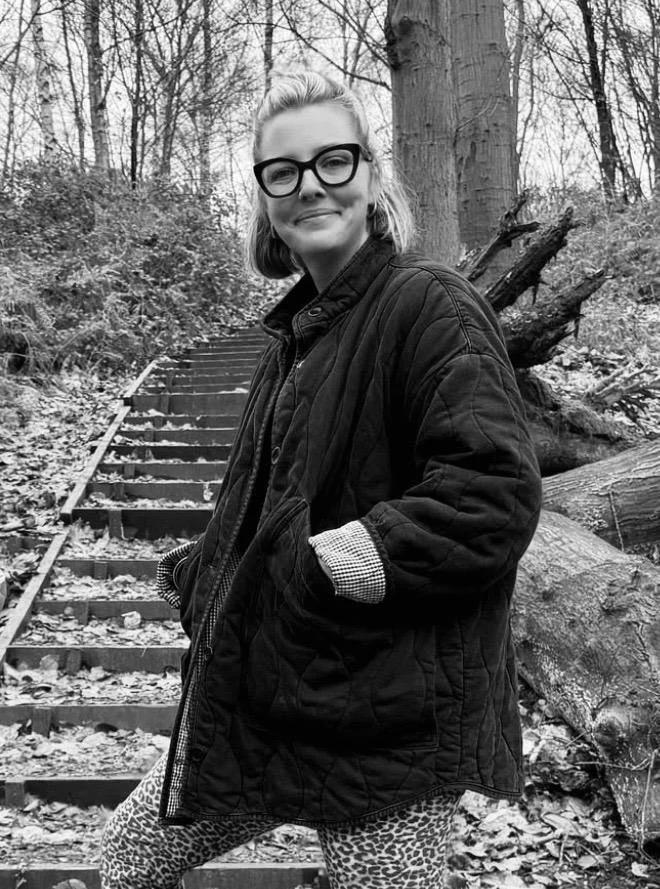 Black and white photo of a young woman with glasses, blonde hair, wearing a quilted jacket and animal print pants, standing on outdoor wooden stairs in a wooded area.