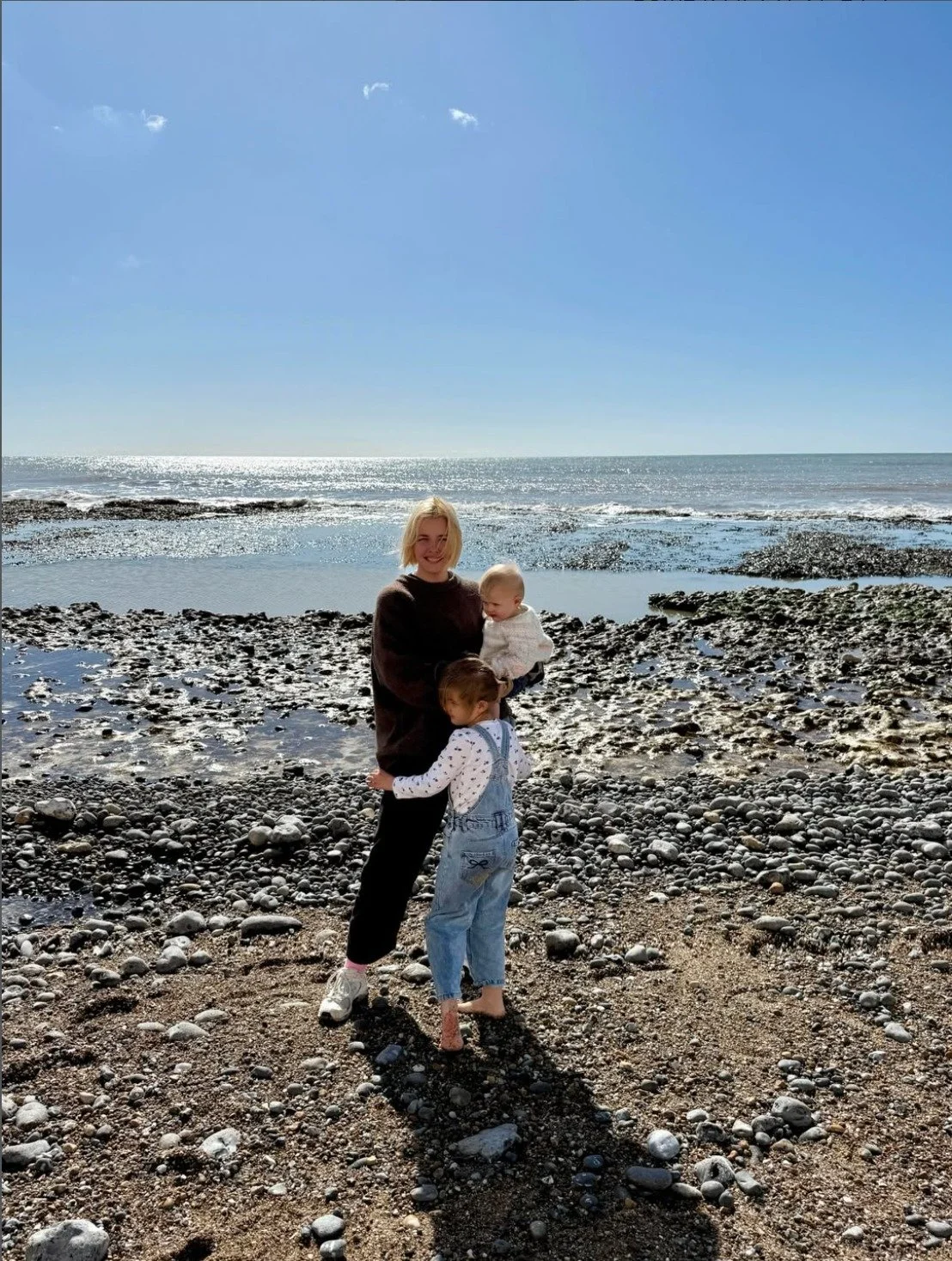 A woman with two children on a rocky beach with the ocean and a clear blue sky in the background.