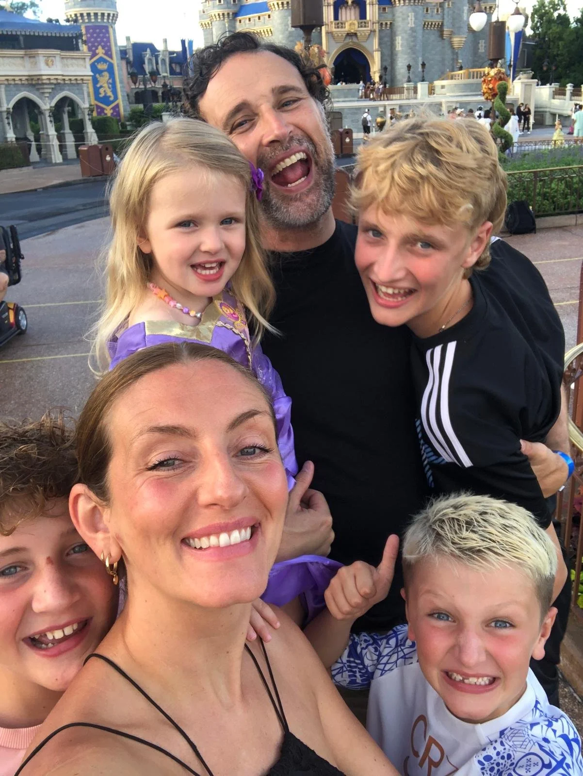 A group of happy people taking a selfie in front of a castle at Disneyland. The group includes three children and three adults, all smiling and enjoying their visit.