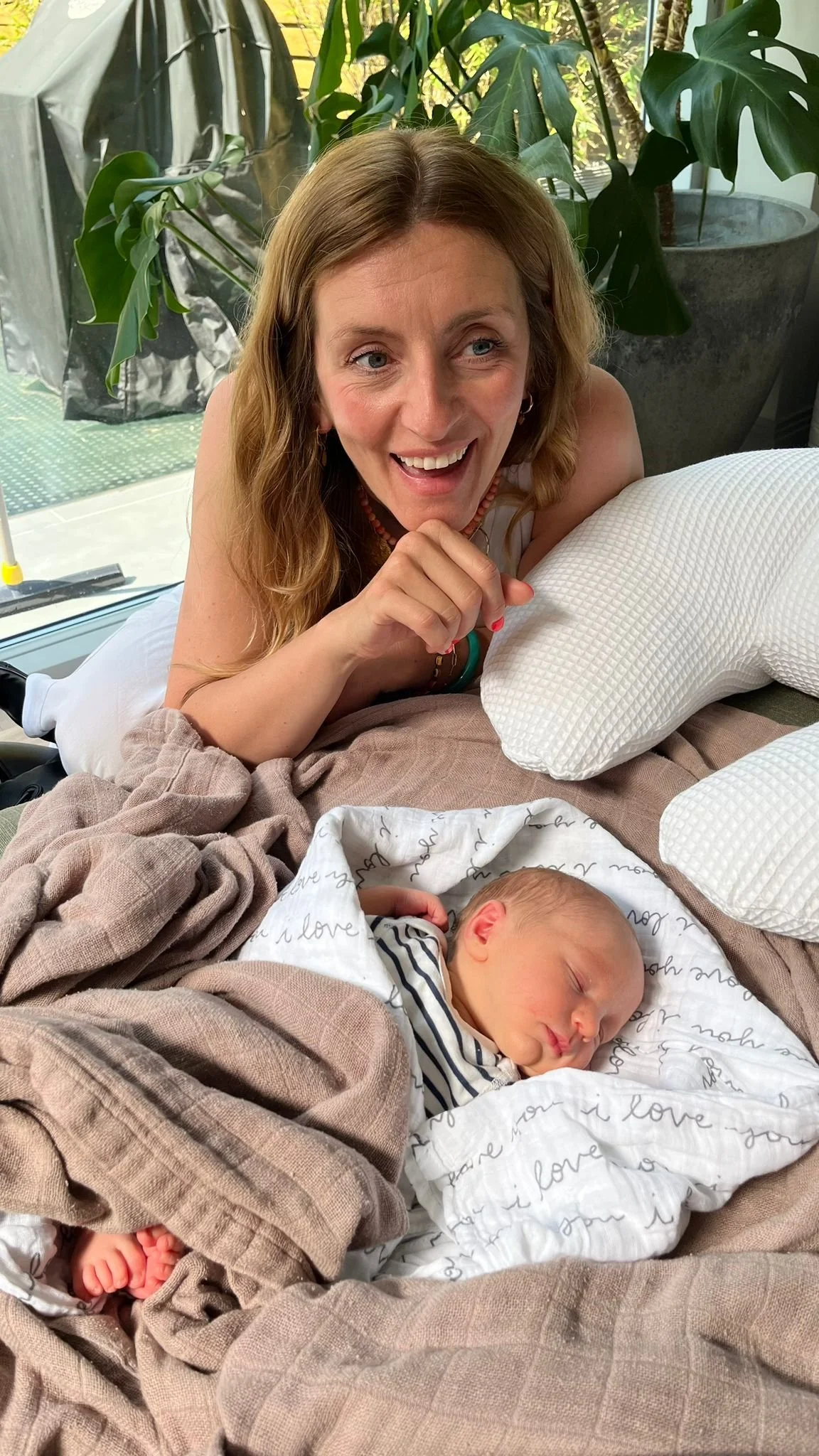A woman with long curly hair smiling and leaning on a pillow, watching a sleeping baby wrapped in blankets, with large green plants and a window in the background.