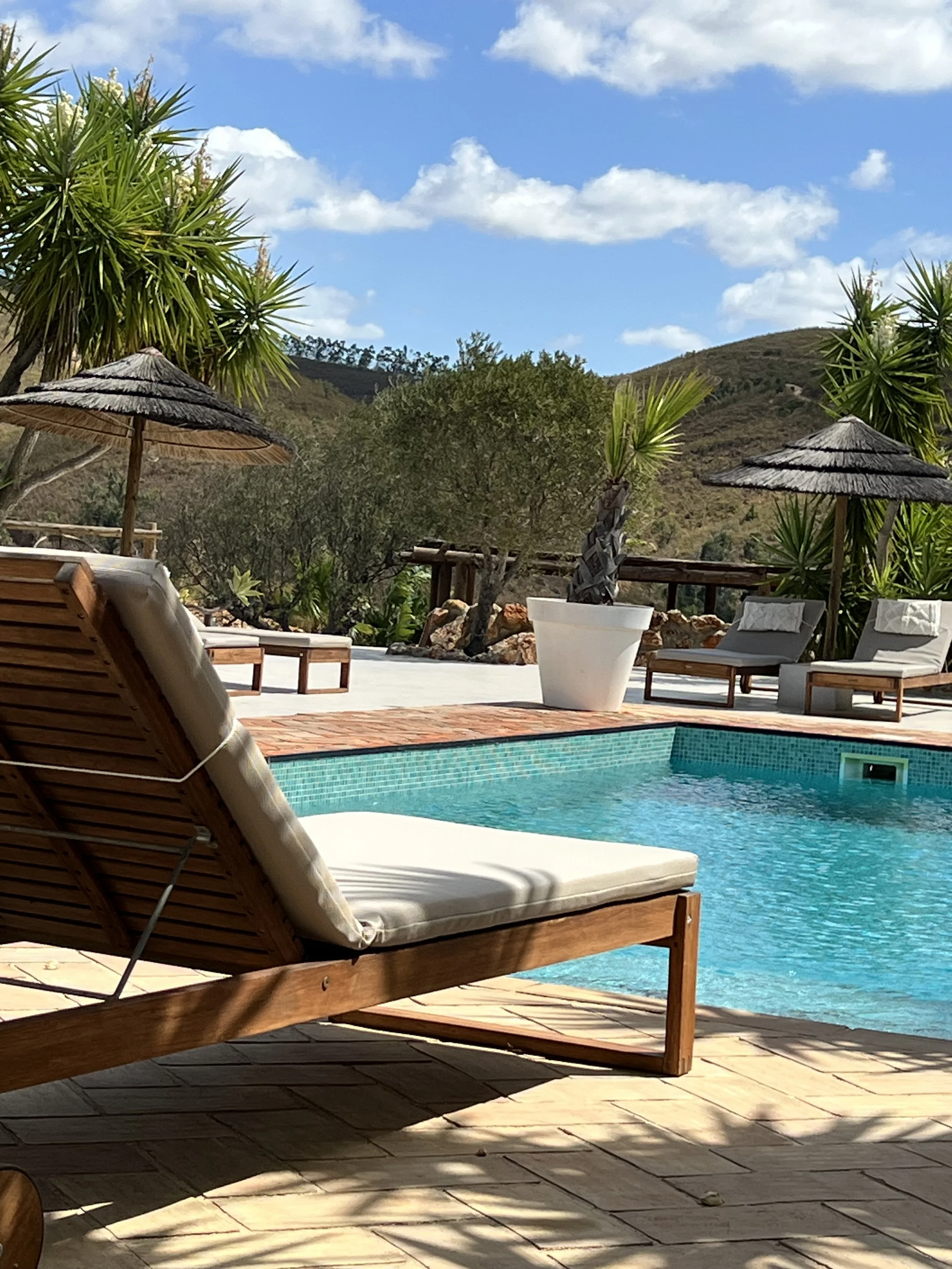 A private outdoor swimming pool with wooden lounge chairs, umbrellas, and tropical plants, set against hills and a partly cloudy sky.