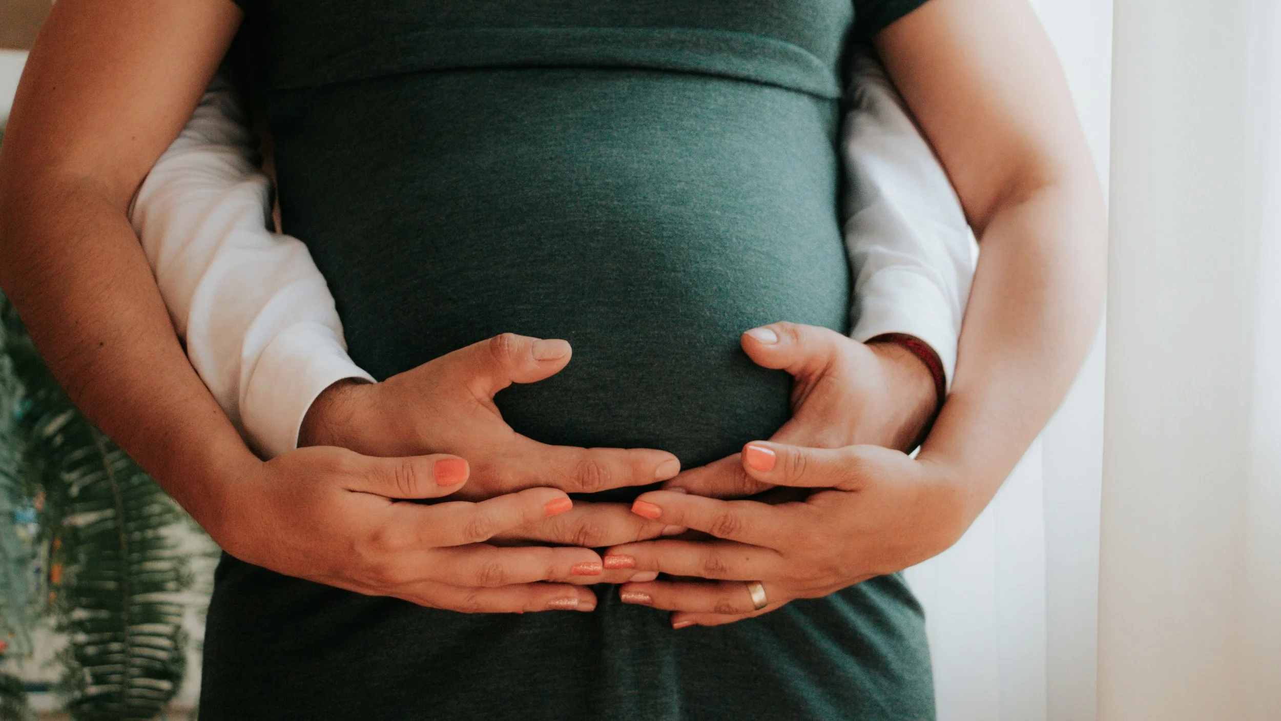 Close-up of a pregnant woman with hands on her belly, with another person's hands gently holding her belly.