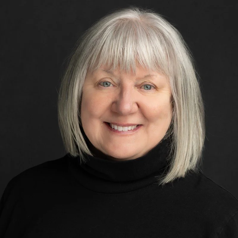 A woman with shoulder-length gray hair smiling, wearing a black turtleneck, against a dark background.