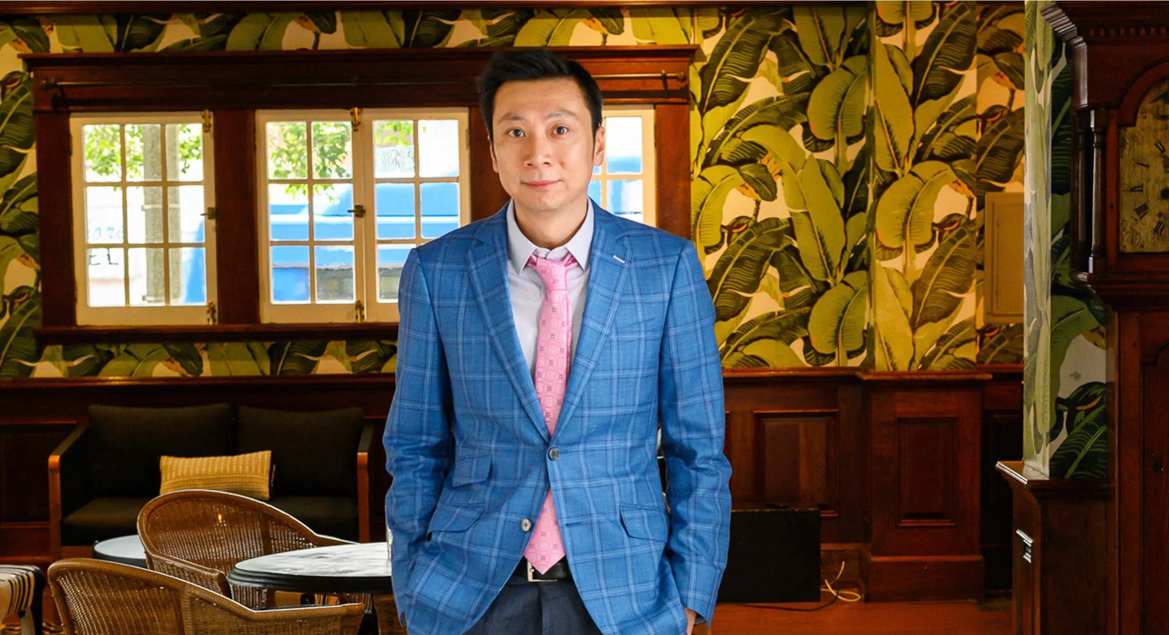 Man standing indoors in a room with tropical leaf wallpaper, wooden window frames, and wicker chairs.
