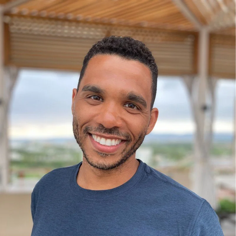 A smiling man with short curly hair, wearing a blue t-shirt, standing outdoors under a wooden structure with a scenic view in the background.