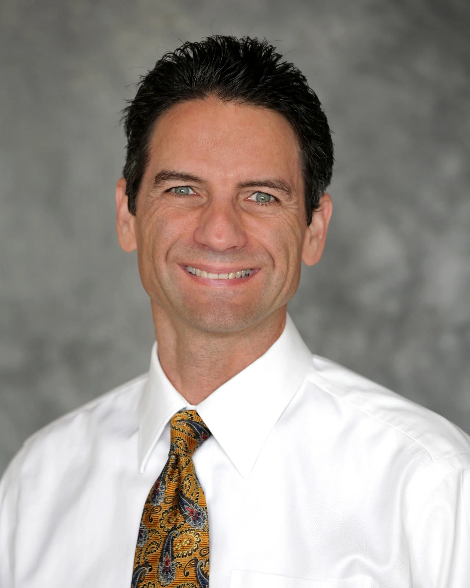 A caucasian man wearing a white shirt and necktie against a gray background, smiling