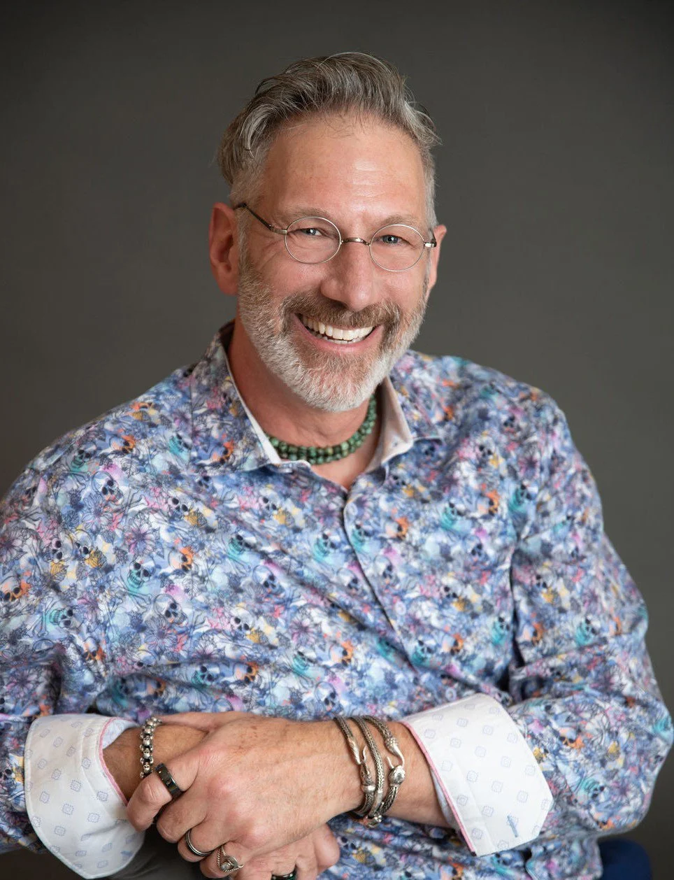 A smiling man with glasses, a beard, and styled gray hair wearing a colorful patterned shirt, a green beaded necklace, and multiple rings and bracelets, with arms crossed.
