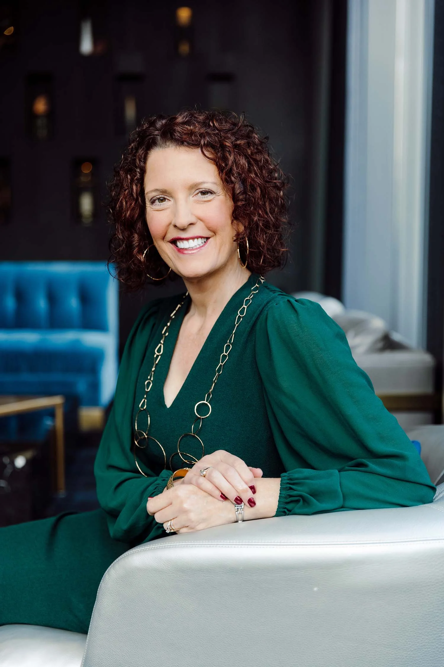 A smiling man with short curly hair, wearing a blue t-shirt, standing outdoors under a wooden structure with a scenic view in the background.
