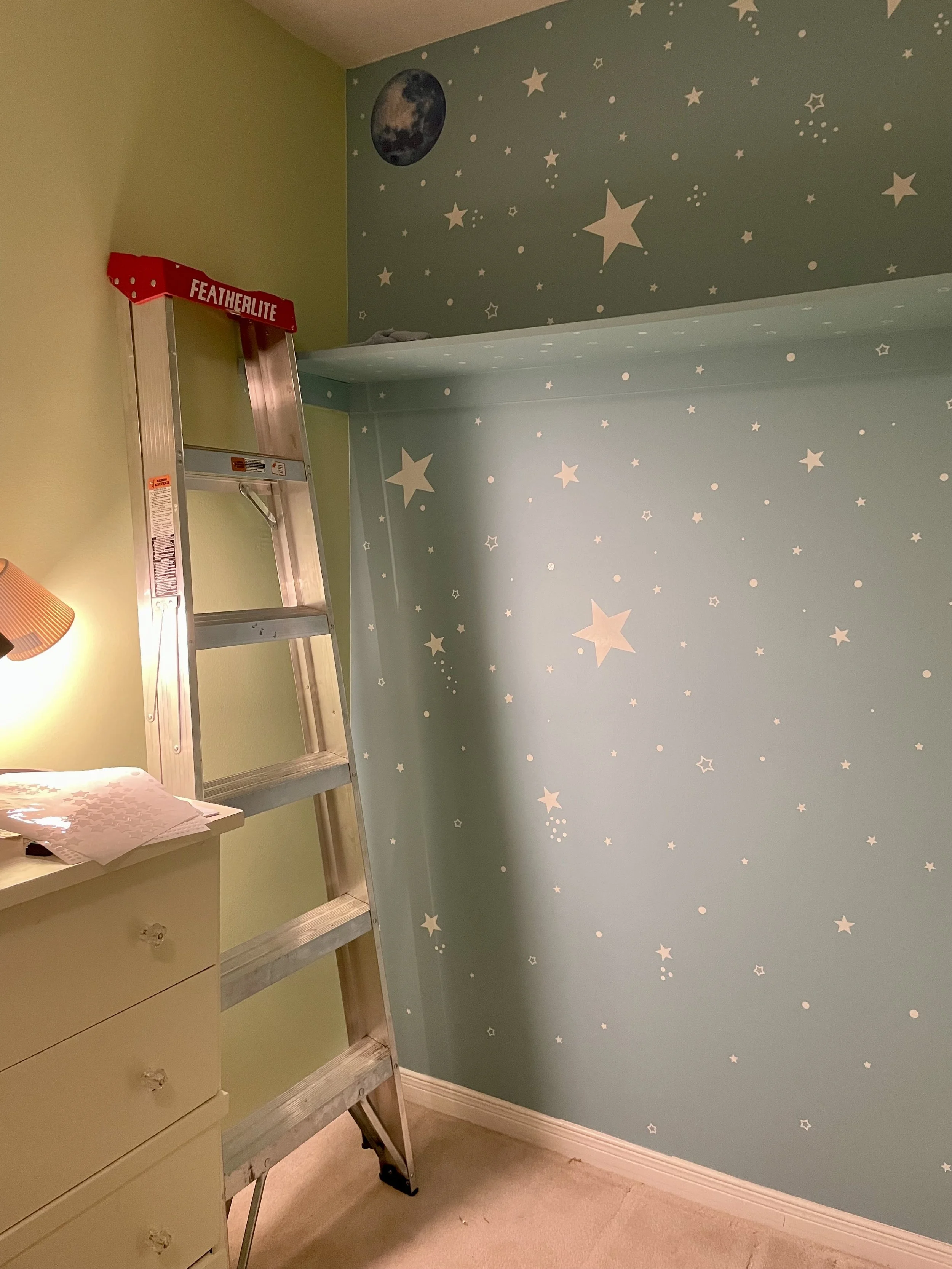A corner of a room with star and moon-themed wall decorations, a silver step ladder with a red "Featherlite" label, a yellow chest of drawers, and a wall-mounted lamp with an orange shade.