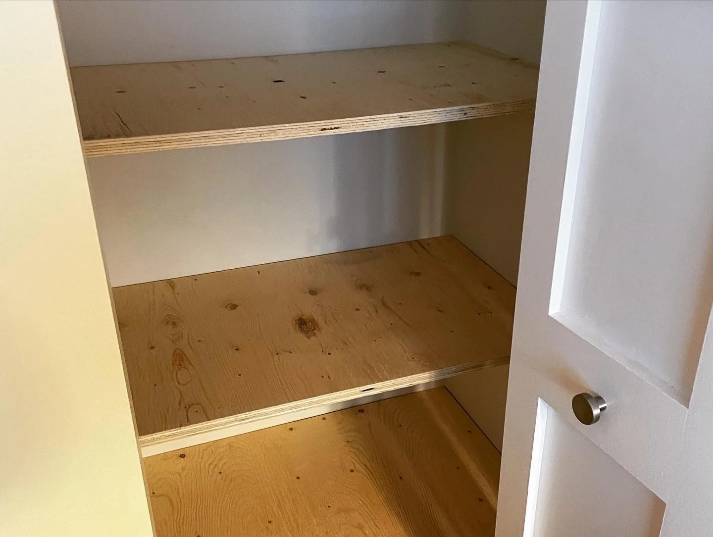 Empty closet with three wooden shelves, white walls, and a white door with a round knob.