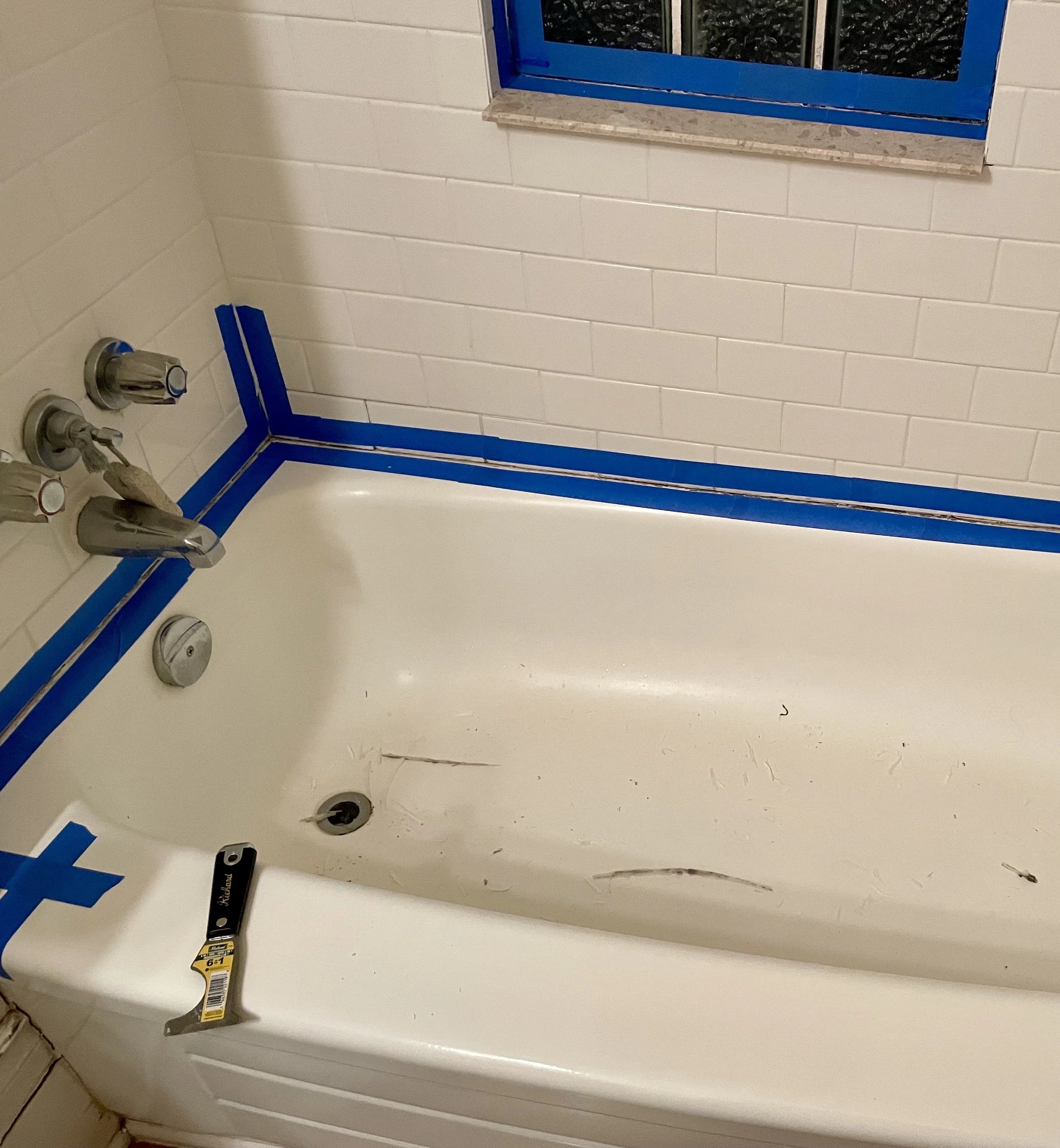 Bathtub with blue painter's tape around edges, repair tools inside, and a window above with a brick sill.