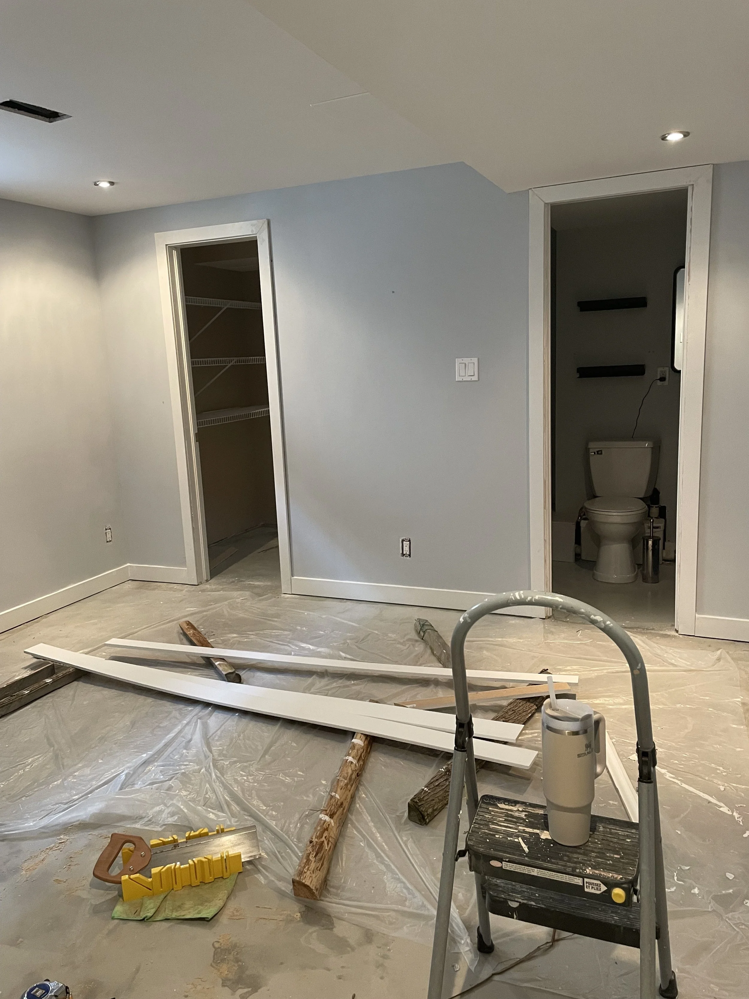 Room under renovation with tools and construction materials, including wood planks, a saw, a step ladder, and a plastic sheet covering the floor. There are two doorways, one leading to a walk-in closet and the other to a small bathroom with a toilet.