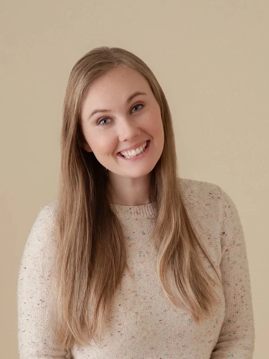 A woman with long, blonde hair, smiling, wearing a beige speckled sweater, standing against a plain beige wall. Photo of Amber Butler, M.Ed, LPC taken by Jessica Barrett Photography.
