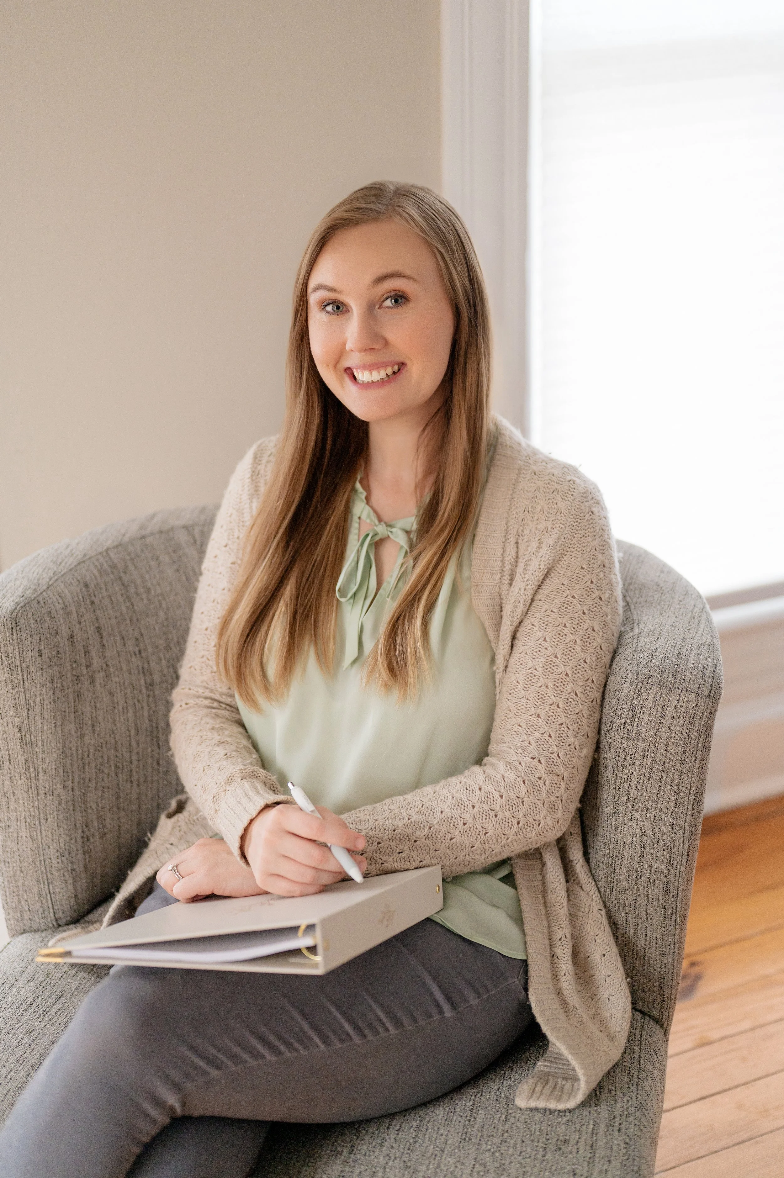 A young woman with long blonde hair, wearing a light green blouse and beige knit cardigan, sitting on a gray sofa with a notepad and pen, smiling at the camera. Photo of Amber Butler, M.Ed, LPC taken by Jessica Barrett Photography.