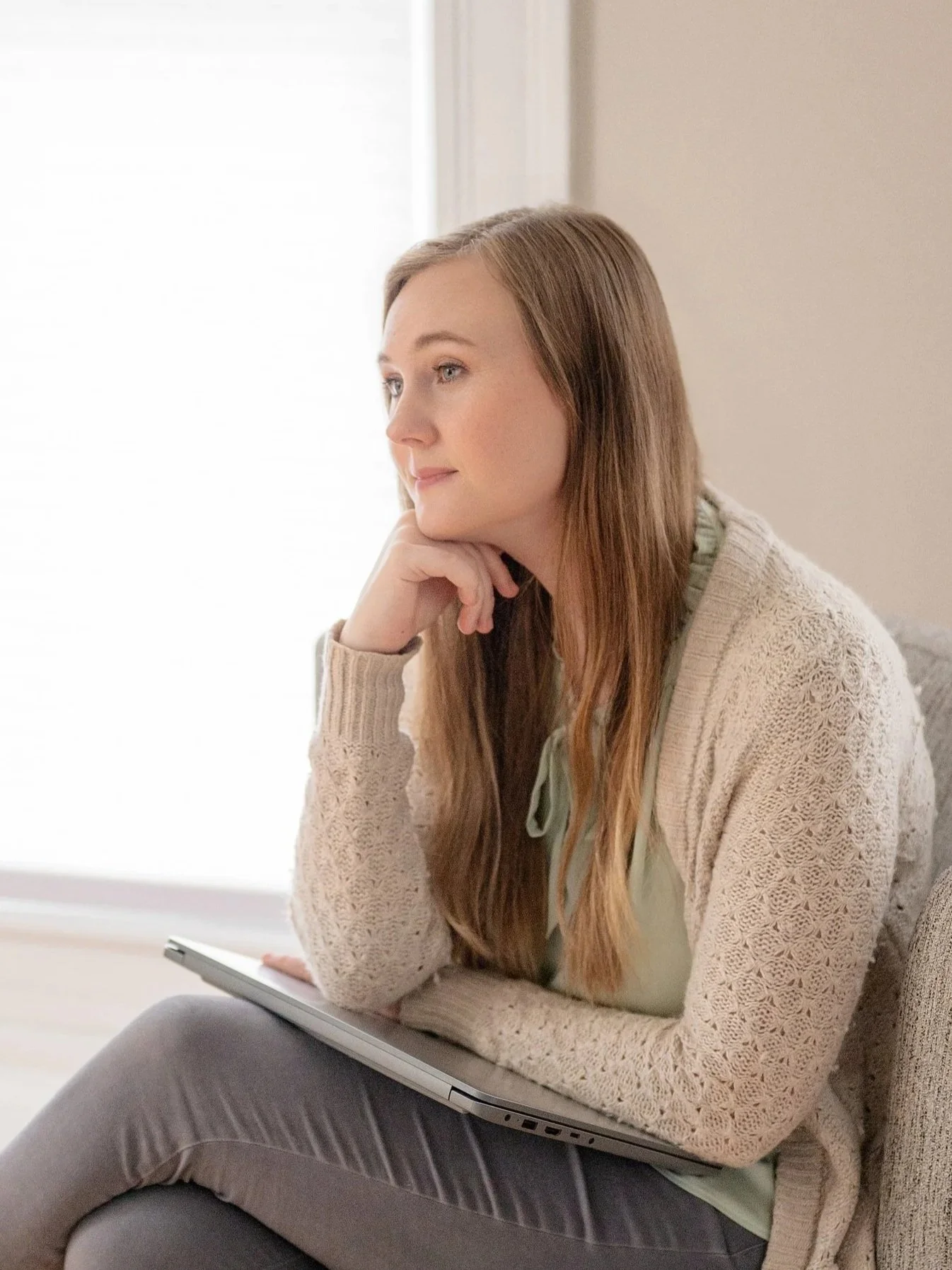 A woman with long blonde hair sitting on a sofa, resting her chin on her hand, looking thoughtfully out a window, with a closed laptop on her lap, in a cozy room. Photo of Amber Butler, M.Ed, LPC taken by Jessica Barrett Photography.