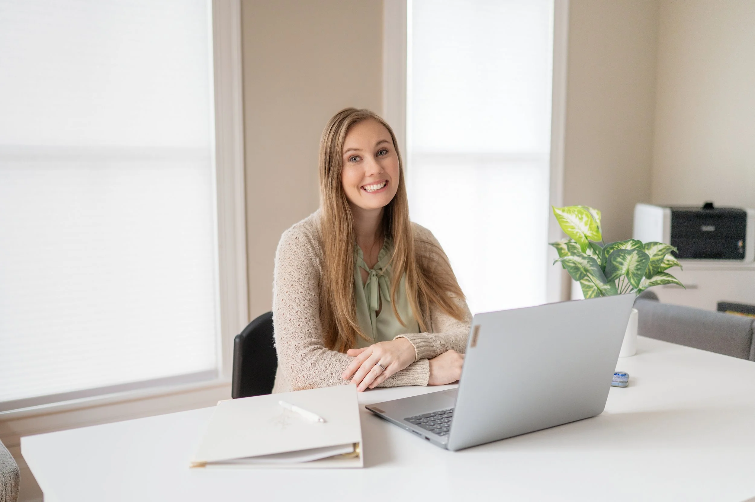 A woman with long blonde hair smiling, sitting at a white desk with a laptop, a potted plant, and a closed folder in a bright room. Photo of Amber Butler, M.Ed, LPC taken by Jessica Barrett Photography.
