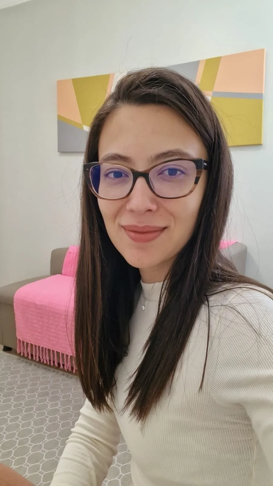 Mulher com cabelos longos e escuros, usando óculos de armação preta, camisa branca, sorrindo, em ambiente com sofá rosa, parede clara e quadro decorativo.