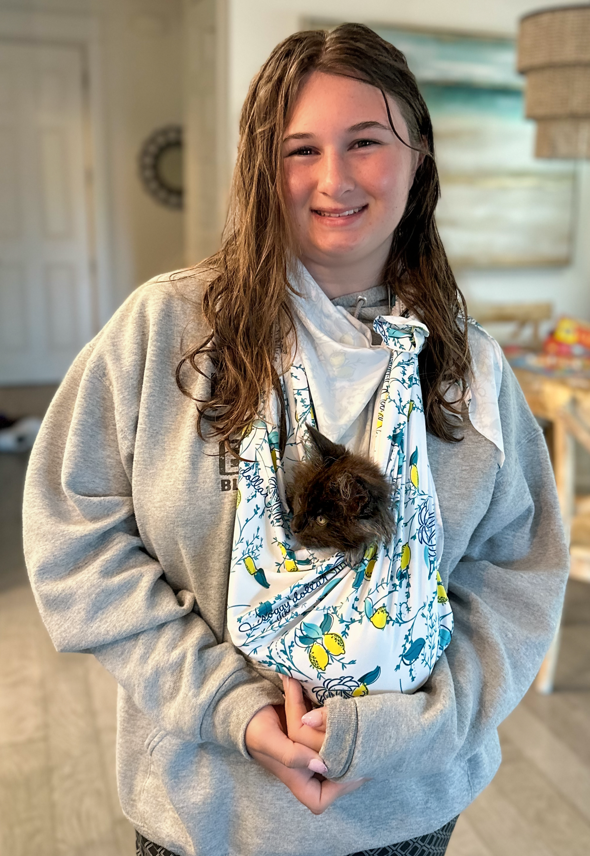 A woman in a grey hoodie holding a black kitten in a patterned bag. The woman is smiling and has wet hair, standing in a cozy home interior.