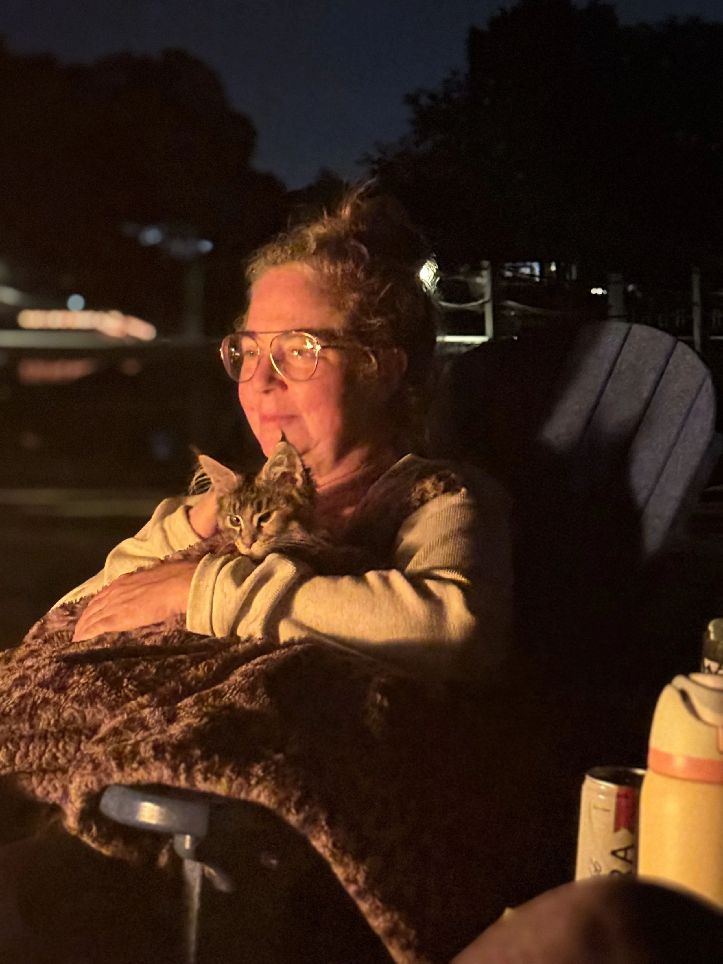 A woman with glasses holding a tabby Maine Coon cat wrapped in a blanket outside at night - Mainecoon Gods LLC