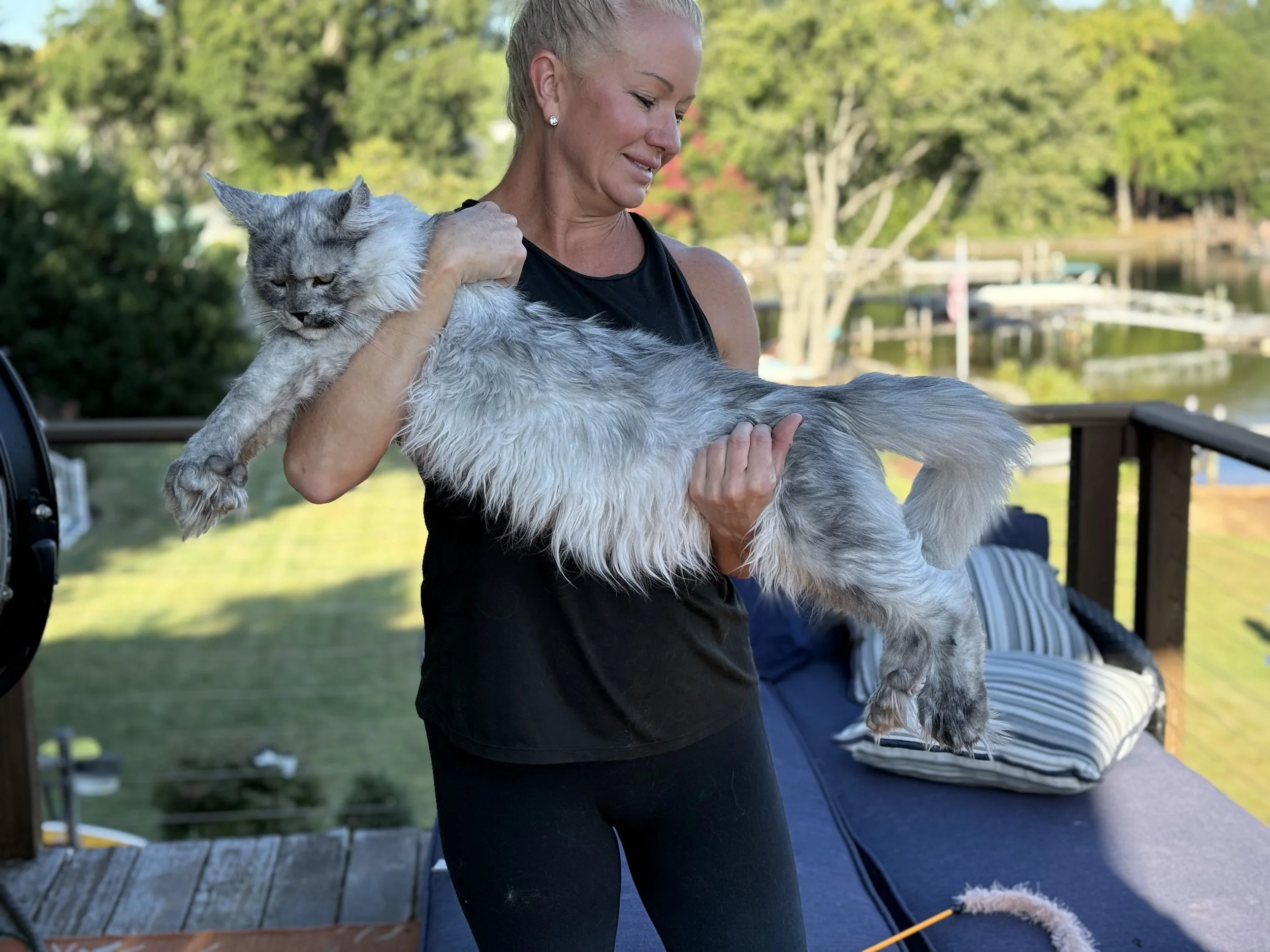 A woman holding a large fluffy gray and white Maine Coon cat outdoors on a deck with a lake, trees, and boats in the background.