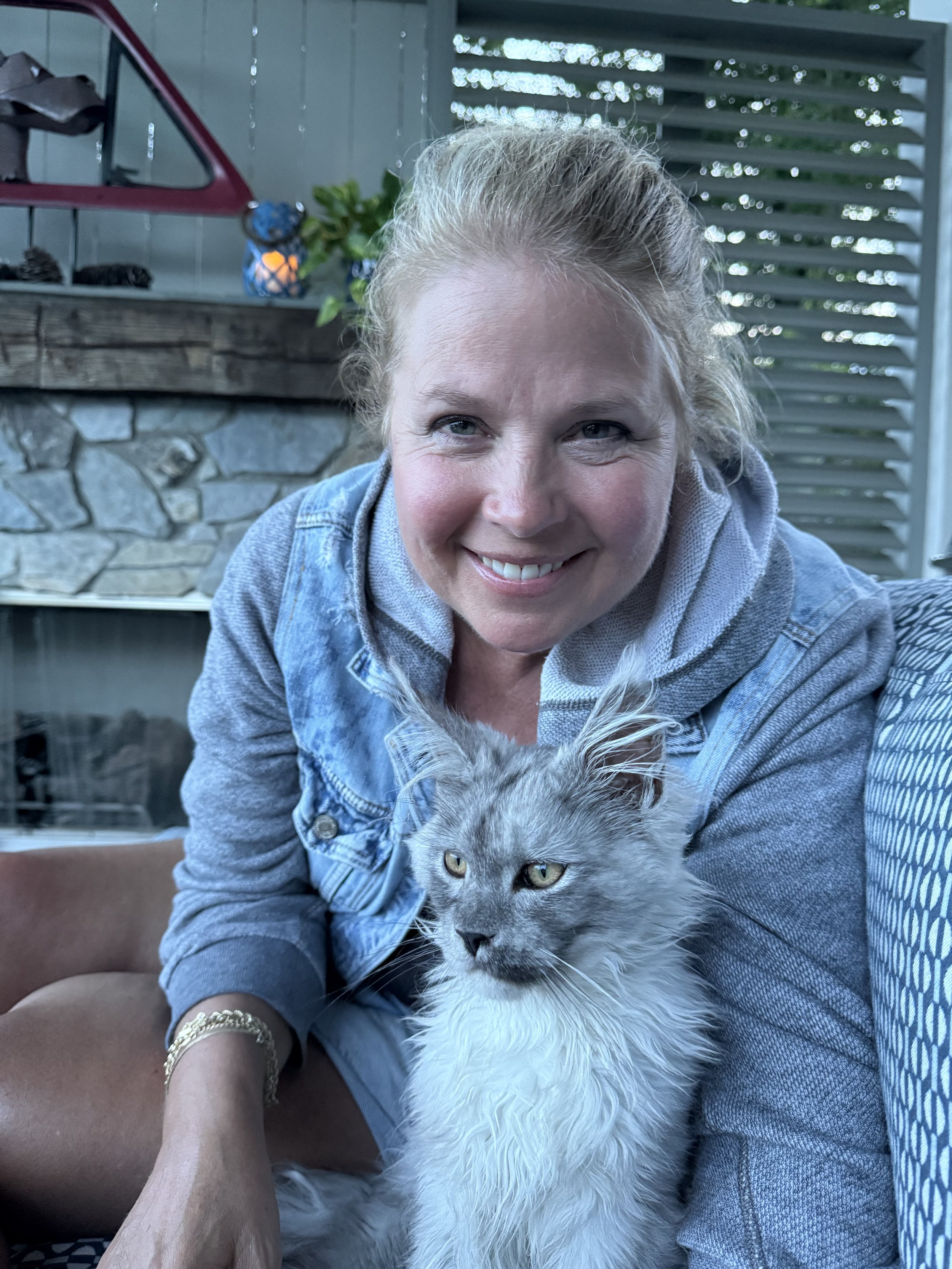 A smiling woman with blonde hair in a ponytail, wearing a denim jacket, sitting outdoors next to a gray and white long-haired Maine Coon cat with piercing yellow eyes - Mainecoon Gods LLC