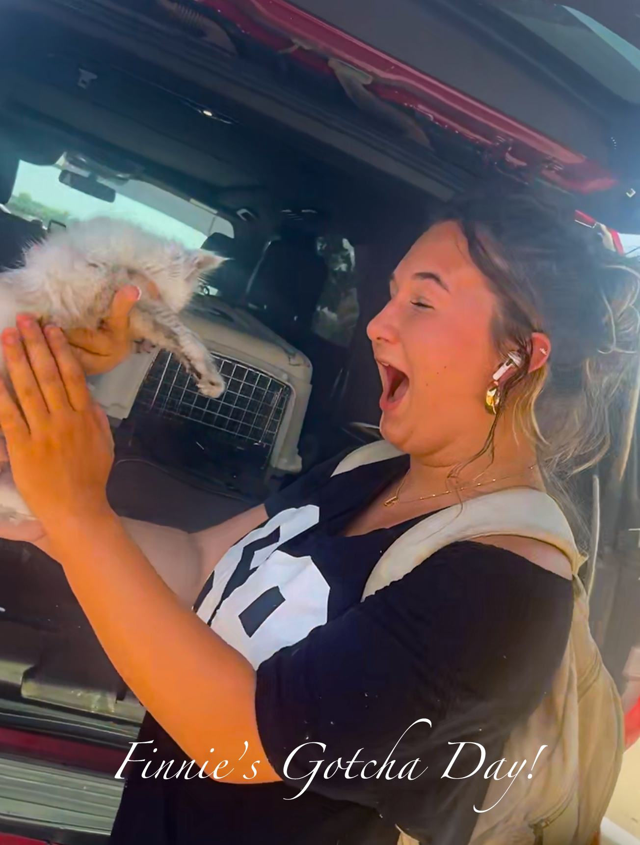 A woman holding a small white kitten next to the open trunk of a vehicle, with a pet carrier inside. The woman is smiling and appears excited. Text overlay reads, 'Finnie's Gotcha Day!'