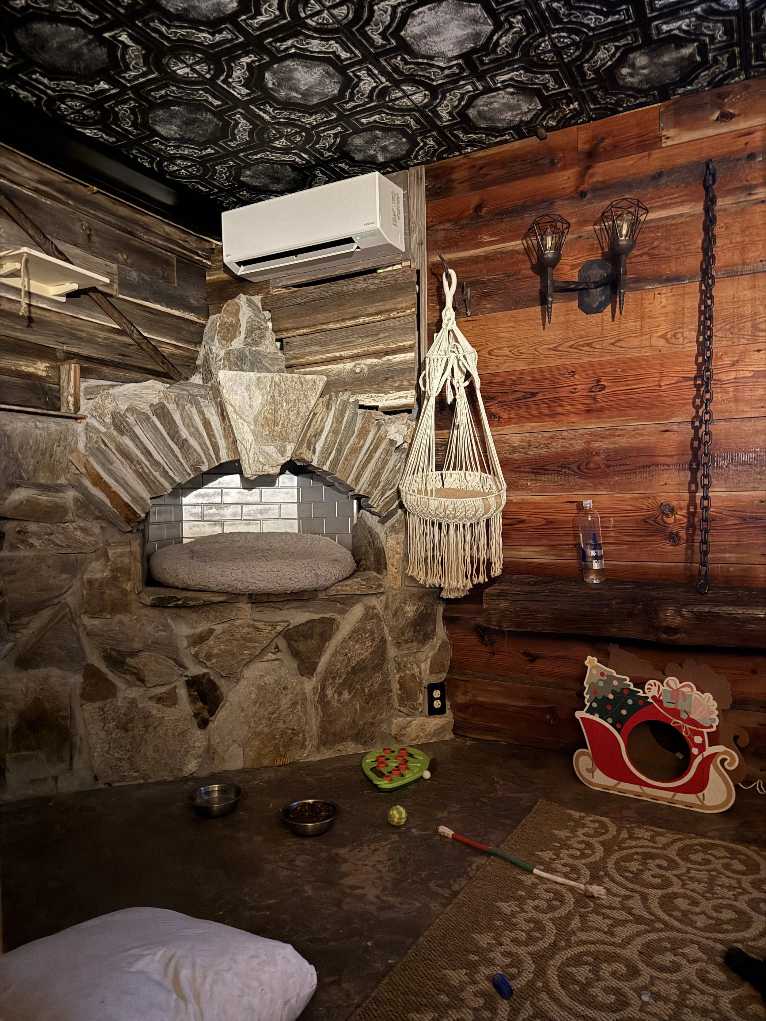 A cozy, rustic room with wooden walls and a stone fireplace with a rounded sitting area. Decorations include a hanging macrame chair, wall-mounted lantern-style lights, a Santa sleigh cutout with a Christmas tree, a water bottle, and scattered toys a