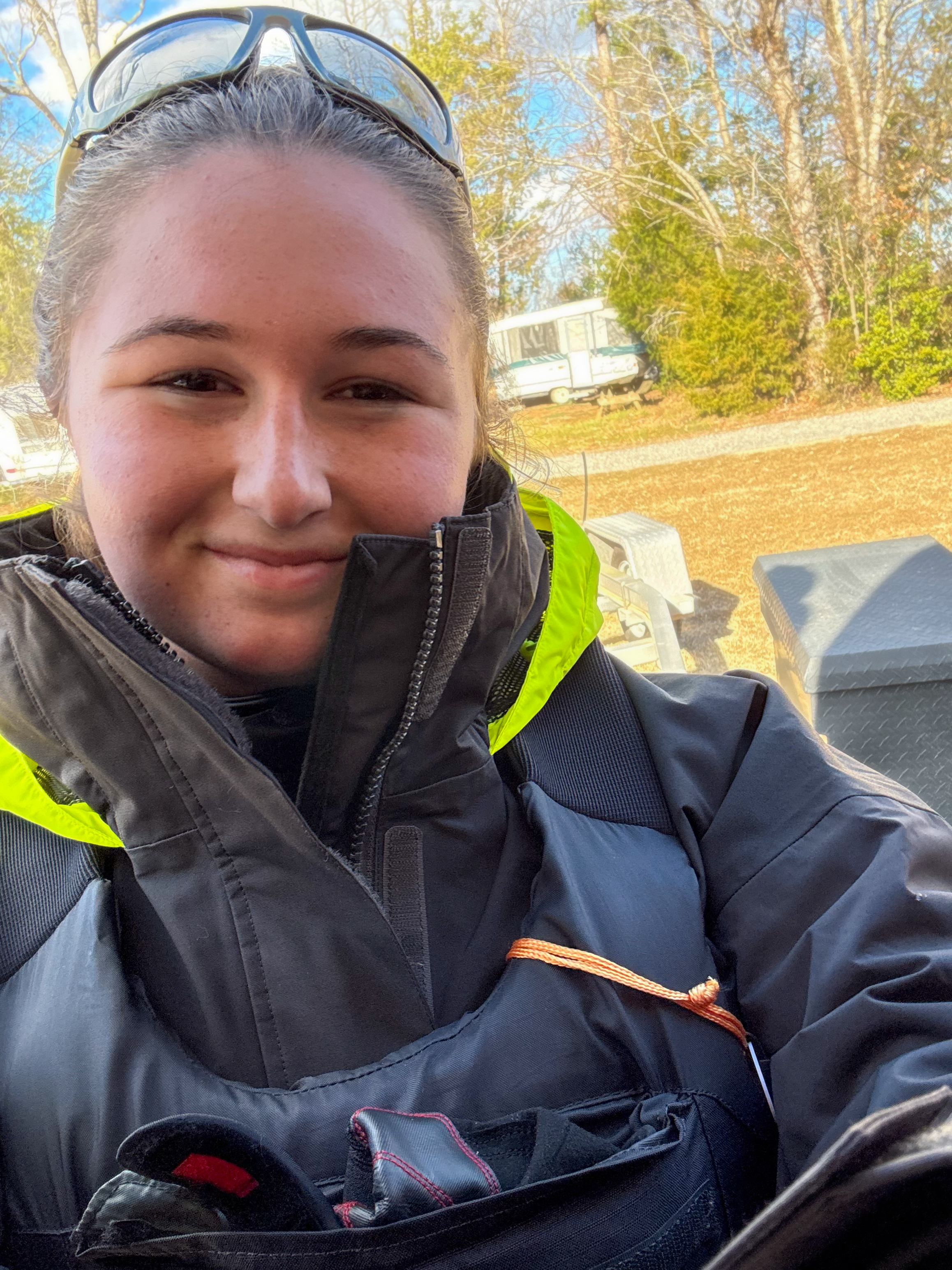 Young woman outdoors in a black and green jacket with sunglasses on her head, smiling at the camera, with trees, a camper, and outdoor equipment visible in the background  - Mainecoon Gods LLC