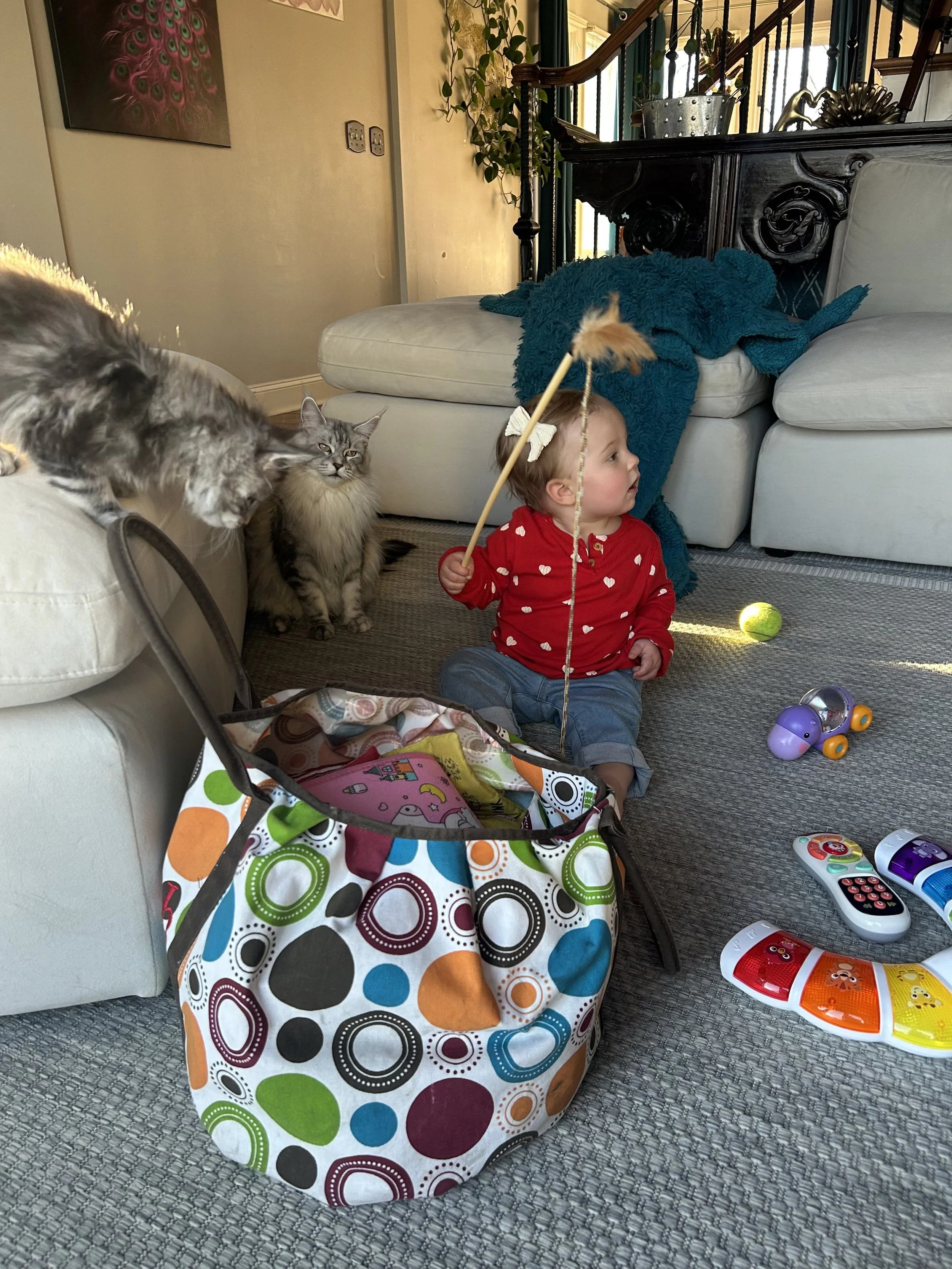 A toddler girl sitting on the living room carpet holding a stick with a feather, surrounded by toys, a cat, and a colorful tote bag.