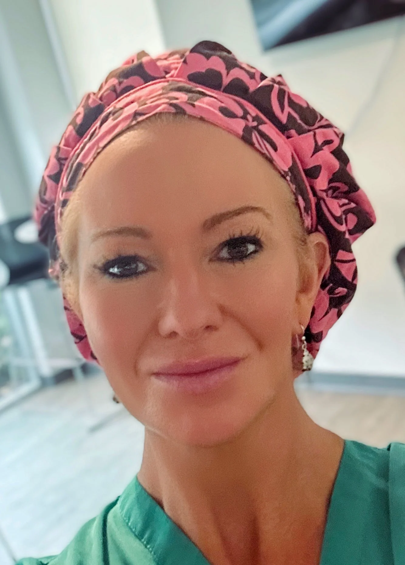 A woman wearing a pink and black patterned scrub cap and green scrubs, smiling at the camera indoors - owner of  Mainecoon Gods LLC