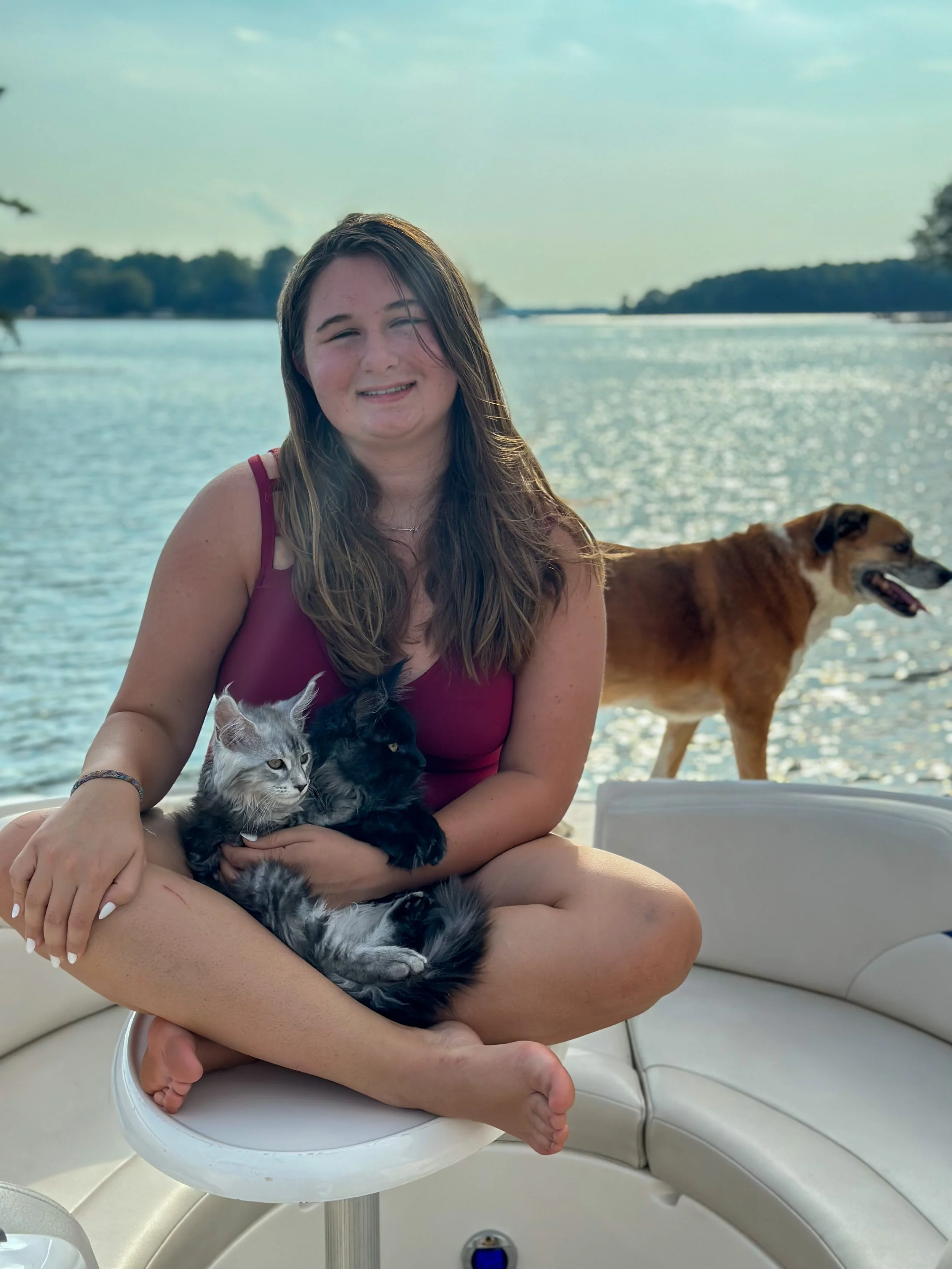 A young woman sitting cross-legged on a boat bench by a lake, holding two cats, with a dog walking nearby. The woman is smiling and wearing a sleeveless purple top, with water and trees in the background.