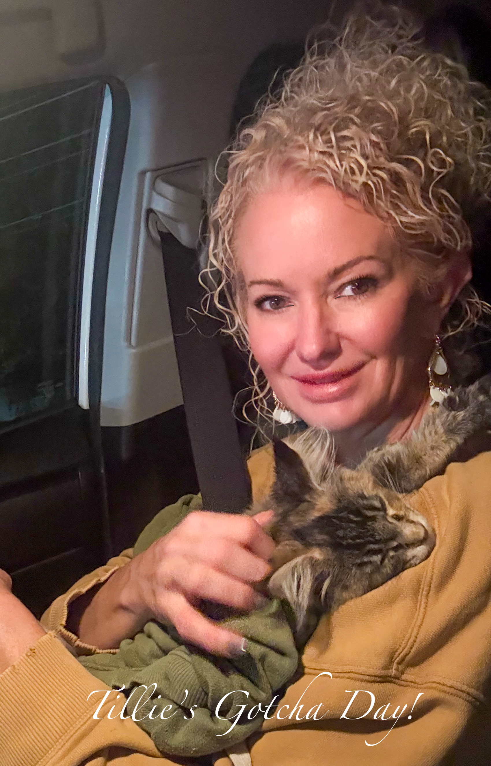 A woman with curly blonde hair holding a sleeping tabby cat, sitting in a car seat at night. The text 'Tiddie's Gotcha Day!' is written at the bottom of the image.
