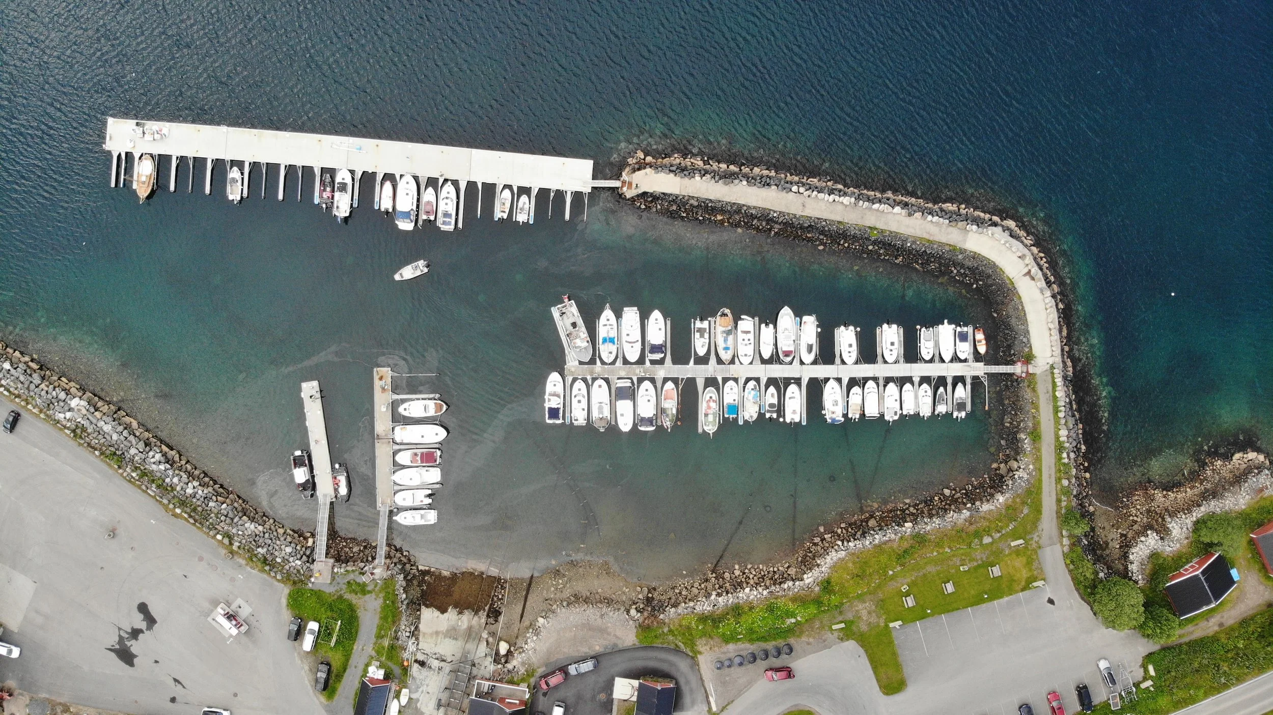 En dronefoto av en småbåthavn med flere båtplasser, steinbrygger og en liten flytebrygge ut i vannet. Det er noen båter i vannet og biler parkert i nærheten av havnen.