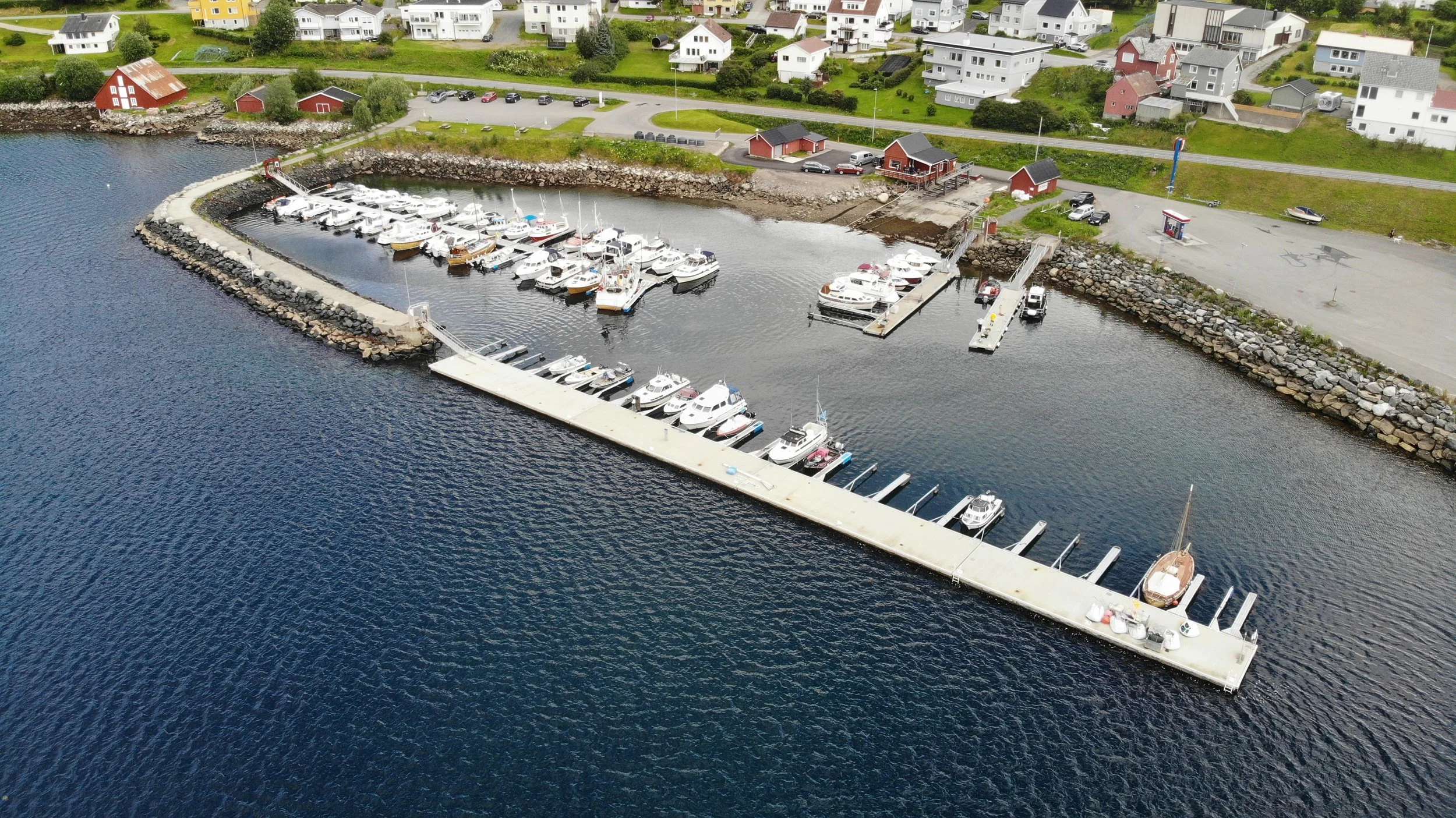 En flyfoto av en liten marina med båtar som ligger til kai, omgitt av en opplyst vei og bolighus på landet.