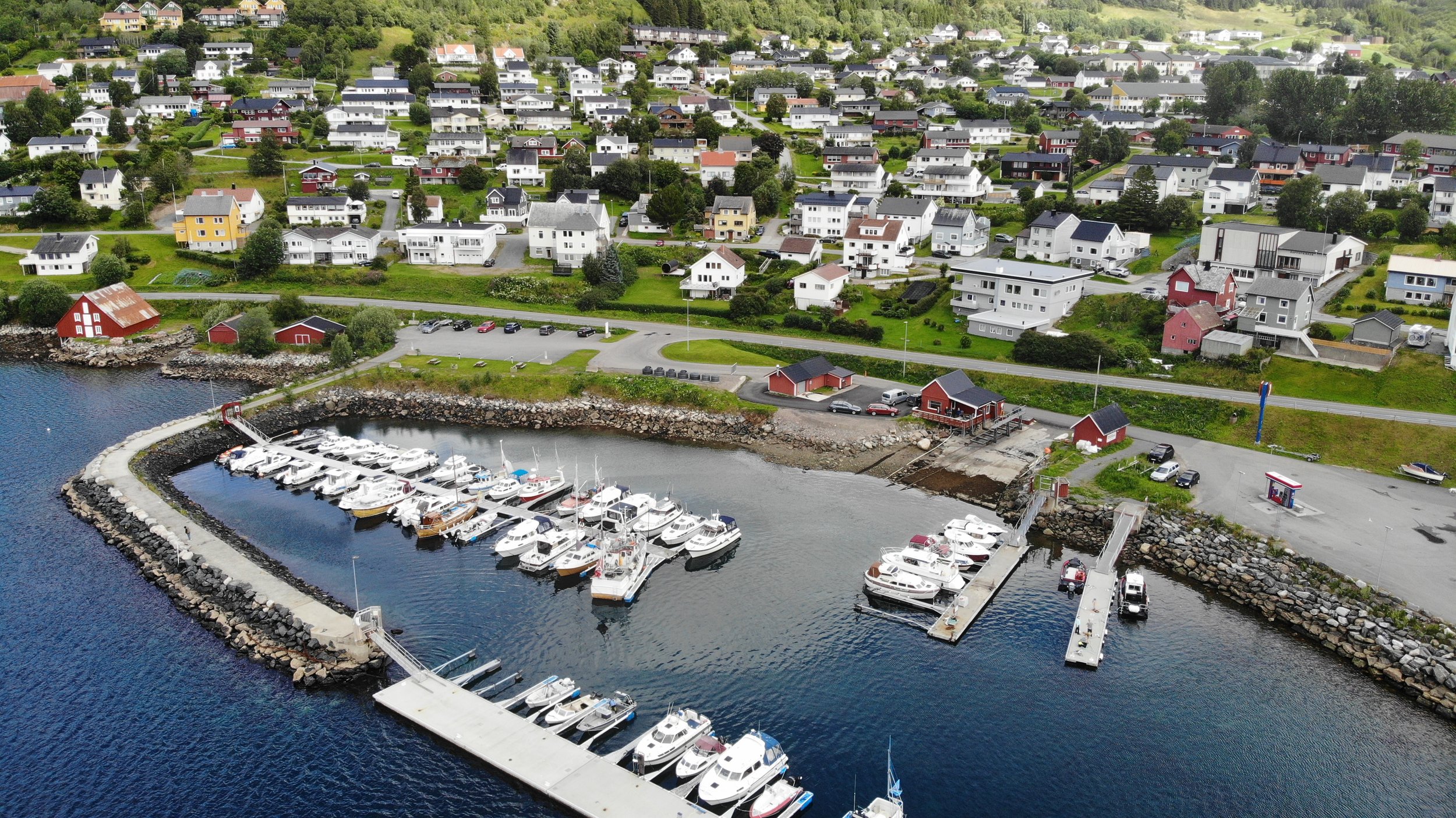 Flyfoto av en liten havn med båter, omgitt av grønne åser og en liten by med mange hus i ulike farger, med skog i bakgrunnen.