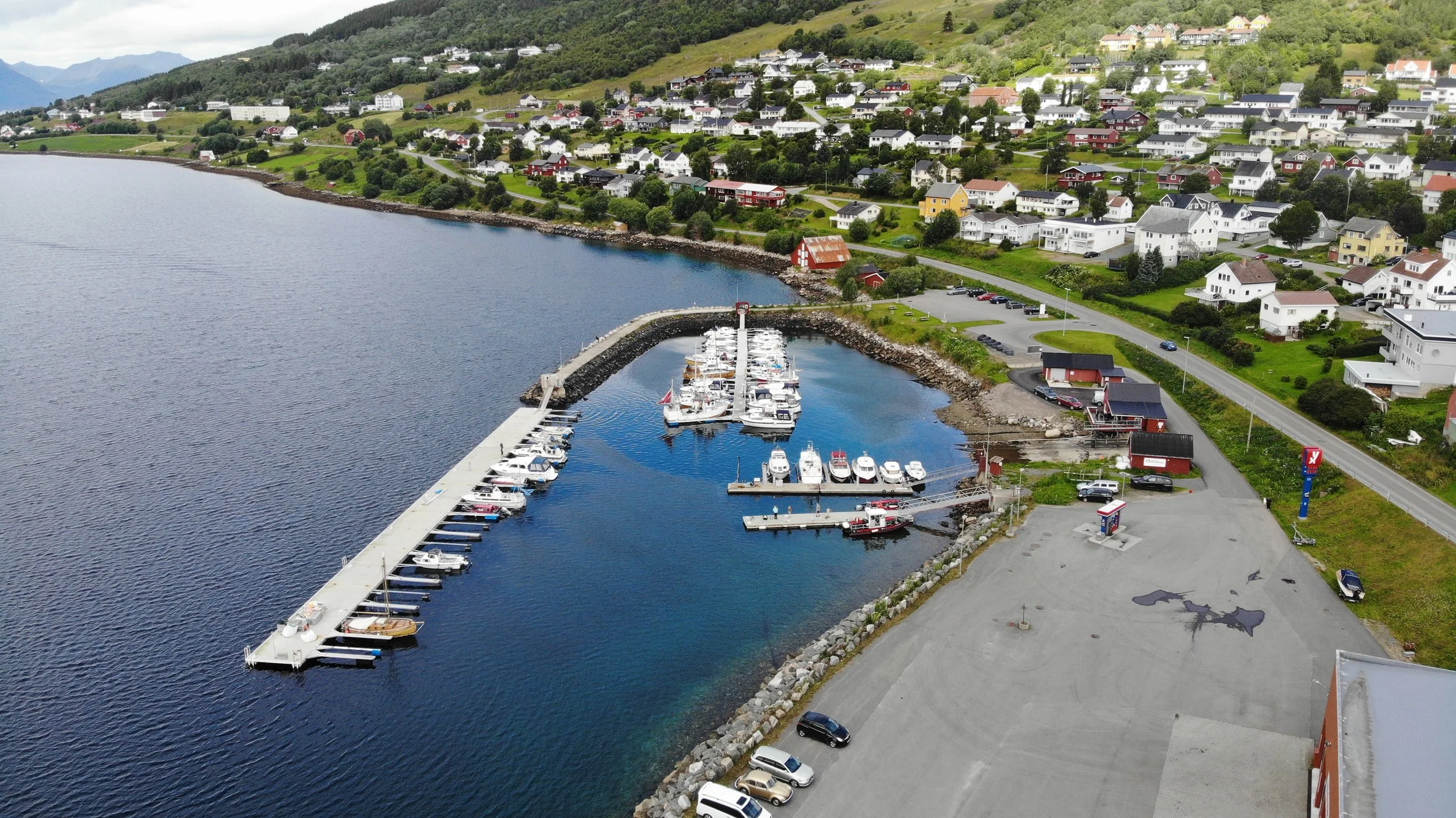 Fjelltopp med små hus spredt over grønt landskap, havn med båter inntill fjorden, parkeringsplass og små bygninger langs vei i en bygder på norsk landsbygd.