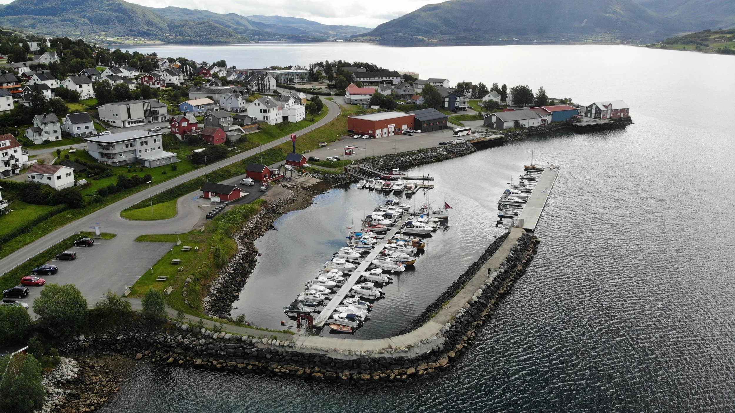 En liten havn med båter og motell med hus i bakgrunnen, omgitt av grønne åser og fjell ved vann.