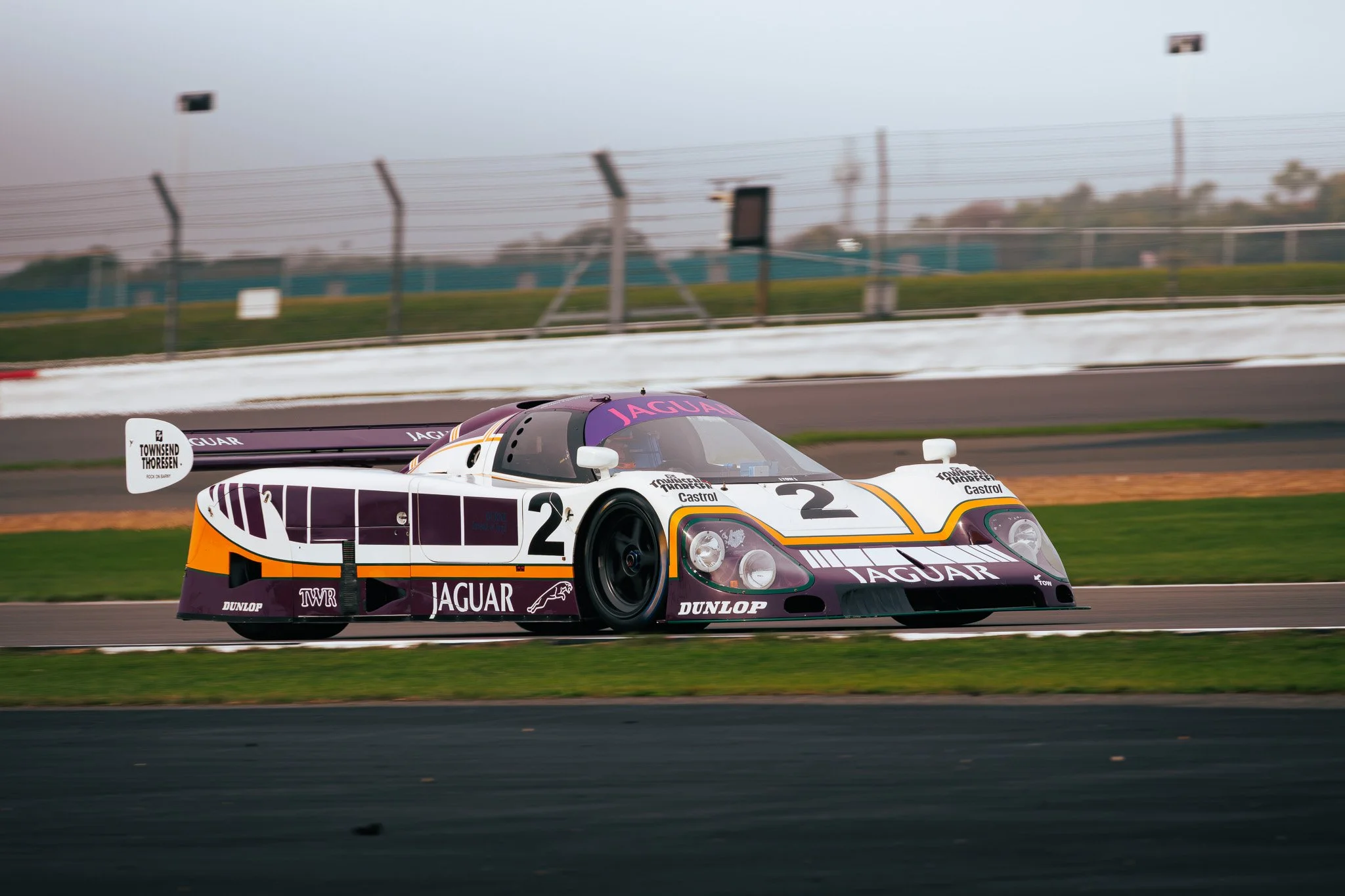 A vintage Jaguar race car on a race track, with a white, purple, and orange livery, and the number 2 on its front and sides.